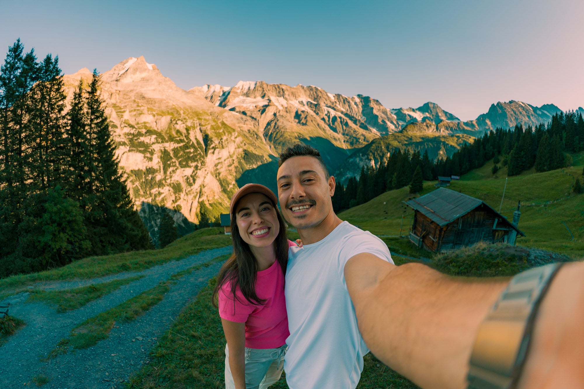 Two people take a selfie with an alpine valley and rugged mountain peaks behind them, photographed by Nick Di Giugno.