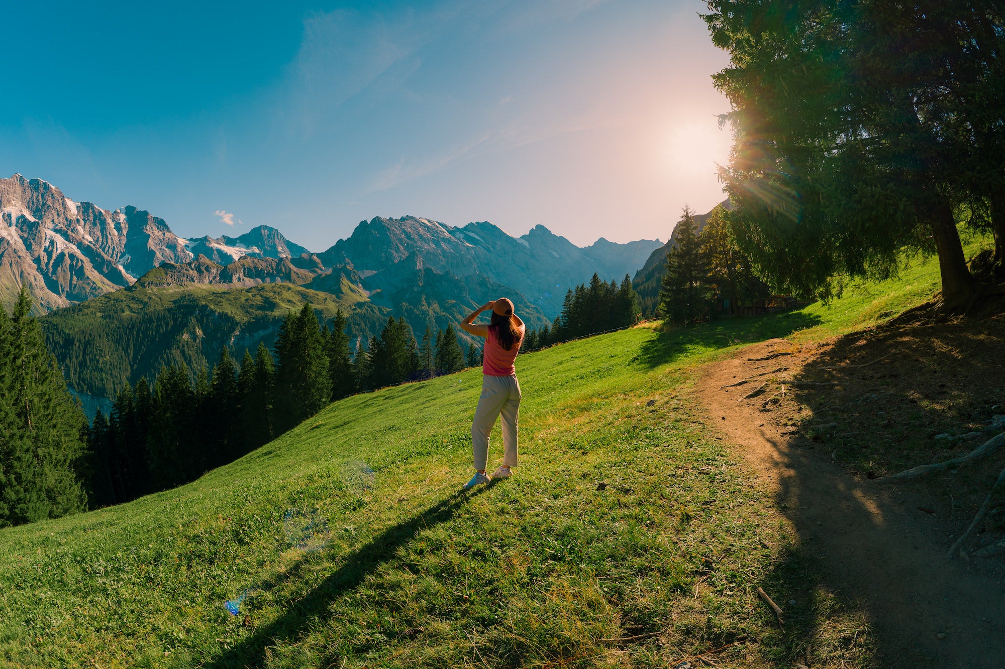 A hiker stands on a grassy mountain slope overlooking a sunlit alpine valley, photographed by Nick Di Giugno.