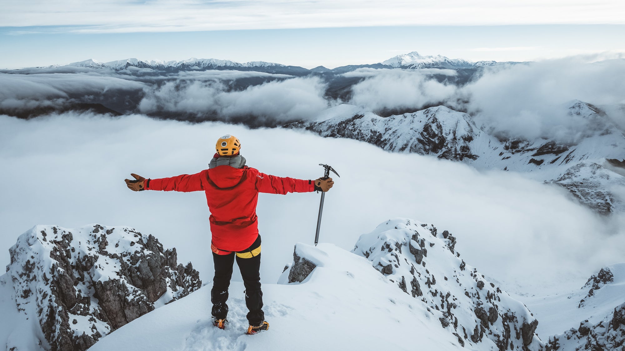 Man in red jacket and yellow helmet standing at the top of a snowy mountain with his hands raised and an ice axe in one hand.
