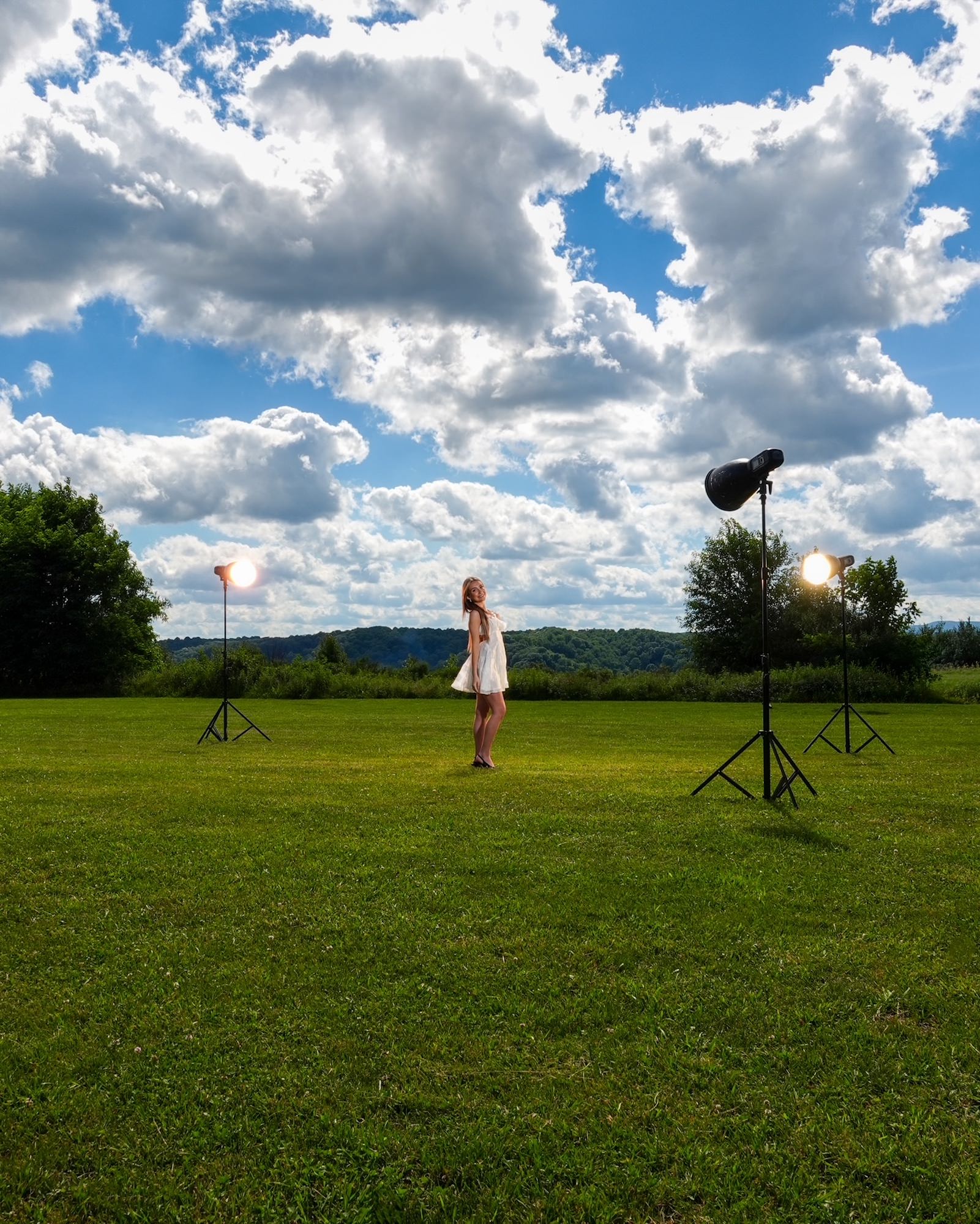 (BTS wide shot) A behind-the-scenes look at an outdoor portrait session: a model in a white dress poses in an open green field flanked by two studio strobes on stands, all set beneath a dramatic cloudy sky &mdash; demonstrating the Sony RX10 V's versatility for