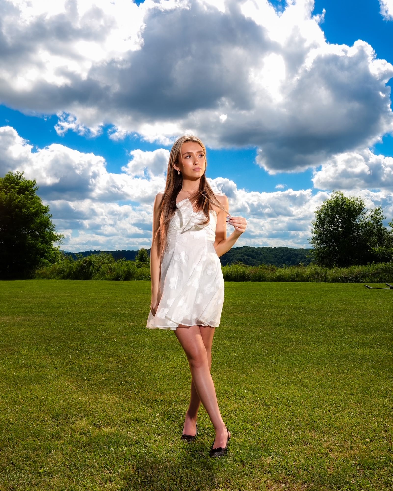 A young woman in a white embroidered mini dress stands poised in a lush green field, looking thoughtfully upward as towering cumulus clouds fill the sky behind her &mdash; shot with the Sony RX10 V.