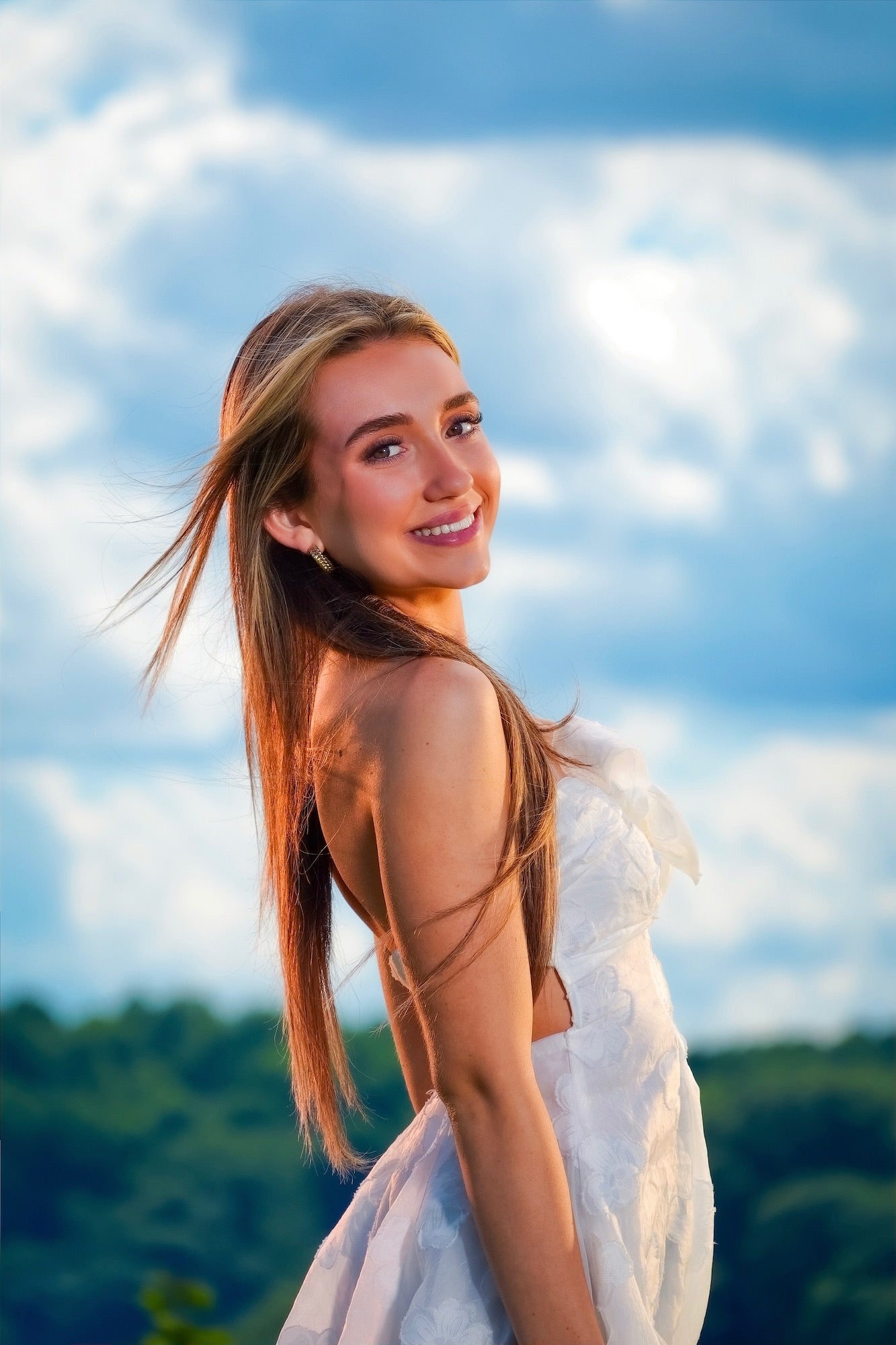 (Model close &mdash; smiling over shoulder) Warm golden light falls across the face of a smiling young woman in a white backless dress as she glances back over her shoulder, a bright cloudy sky and green hills creating a dreamy outdoor portrait backdrop.