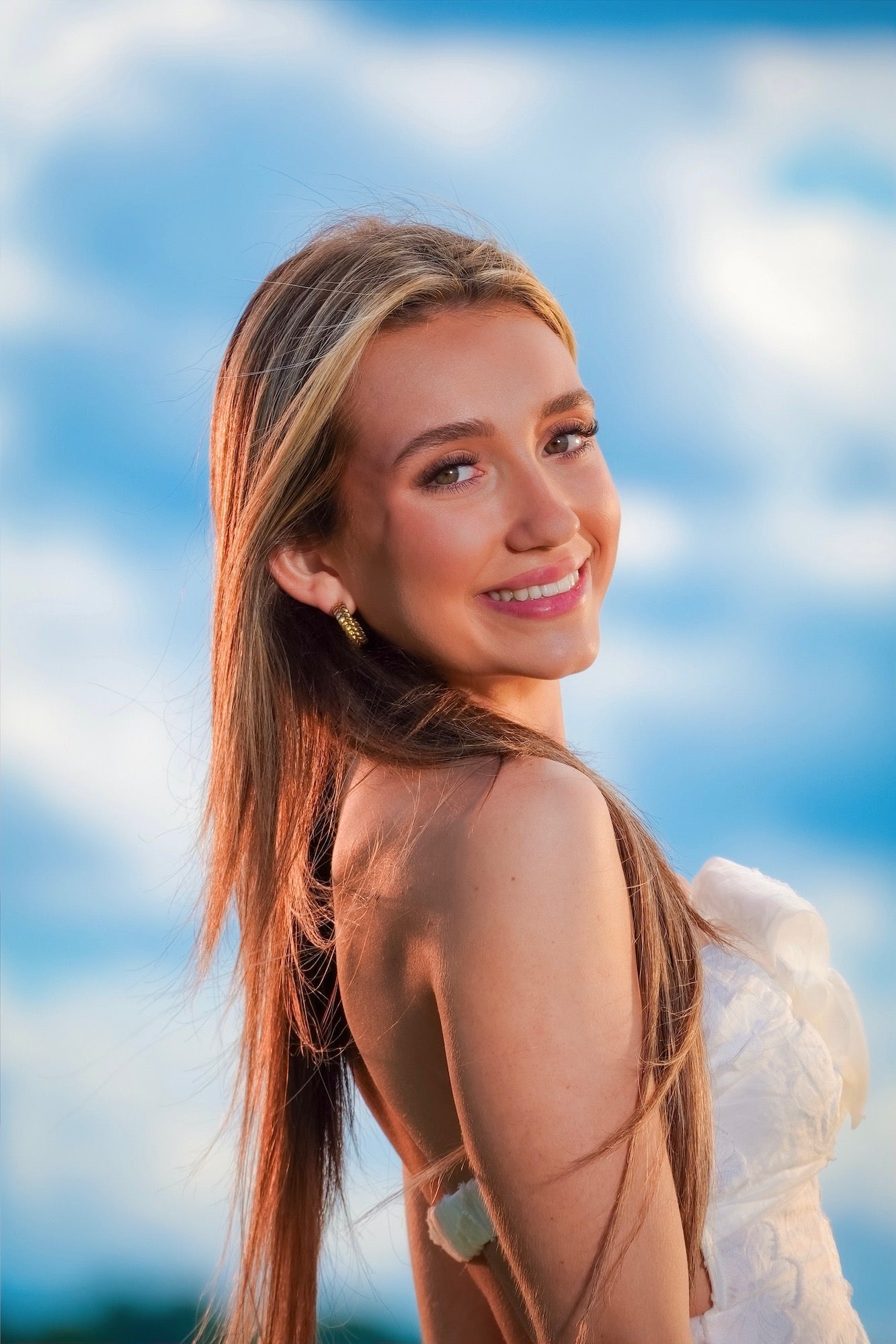 (Model &mdash; tight portrait smiling) A radiant close-up portrait of a young woman smiling over her shoulder, her long hair glowing in warm afternoon light against a soft blue sky filled with white clouds &mdash; shot with the Sony RX10 V.