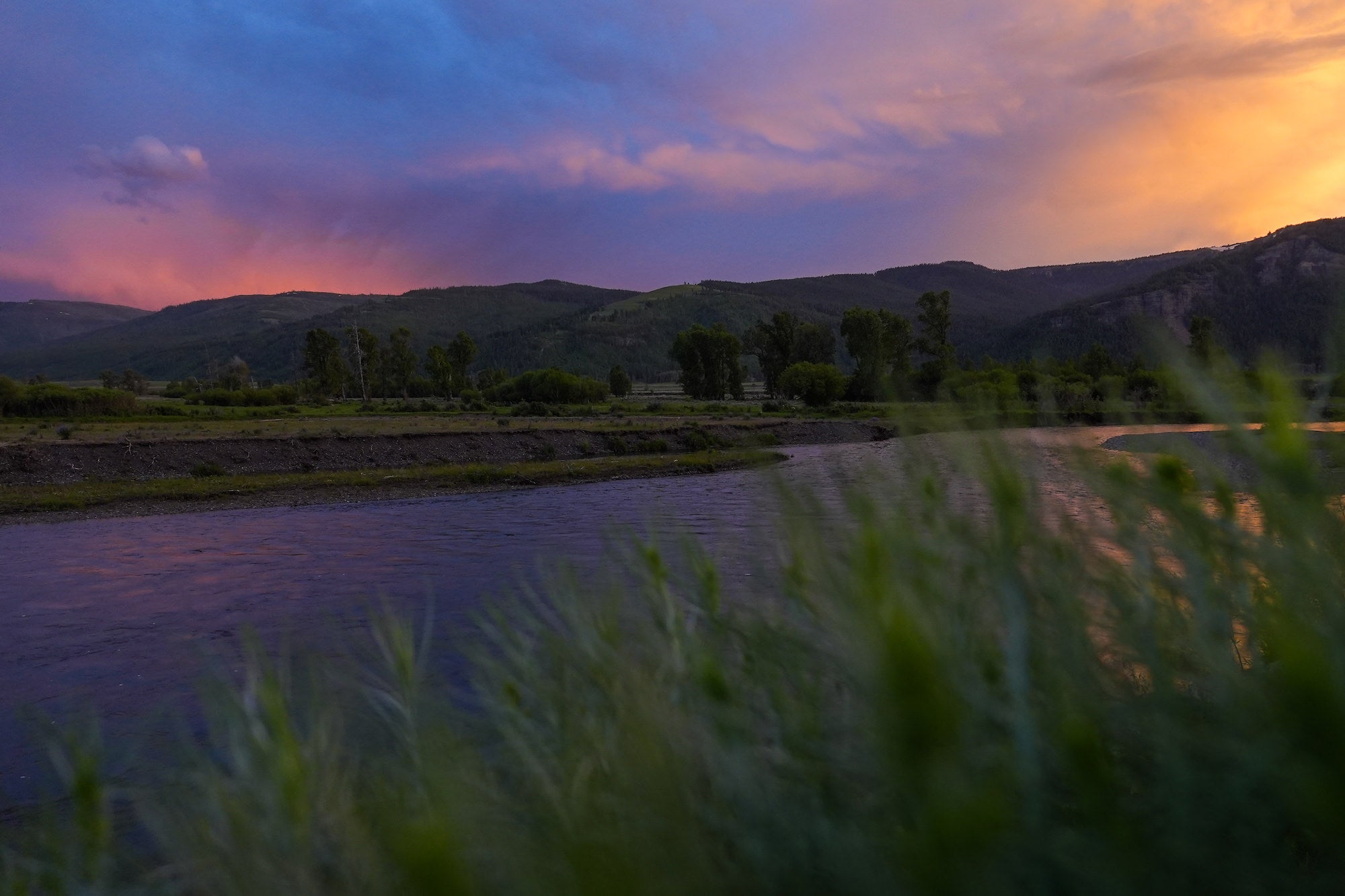 A vivid sunset paints the sky over a mountain river, with blurred wildgrass in the foreground, photographed with the Sony RX10 V by Dani Hamm.