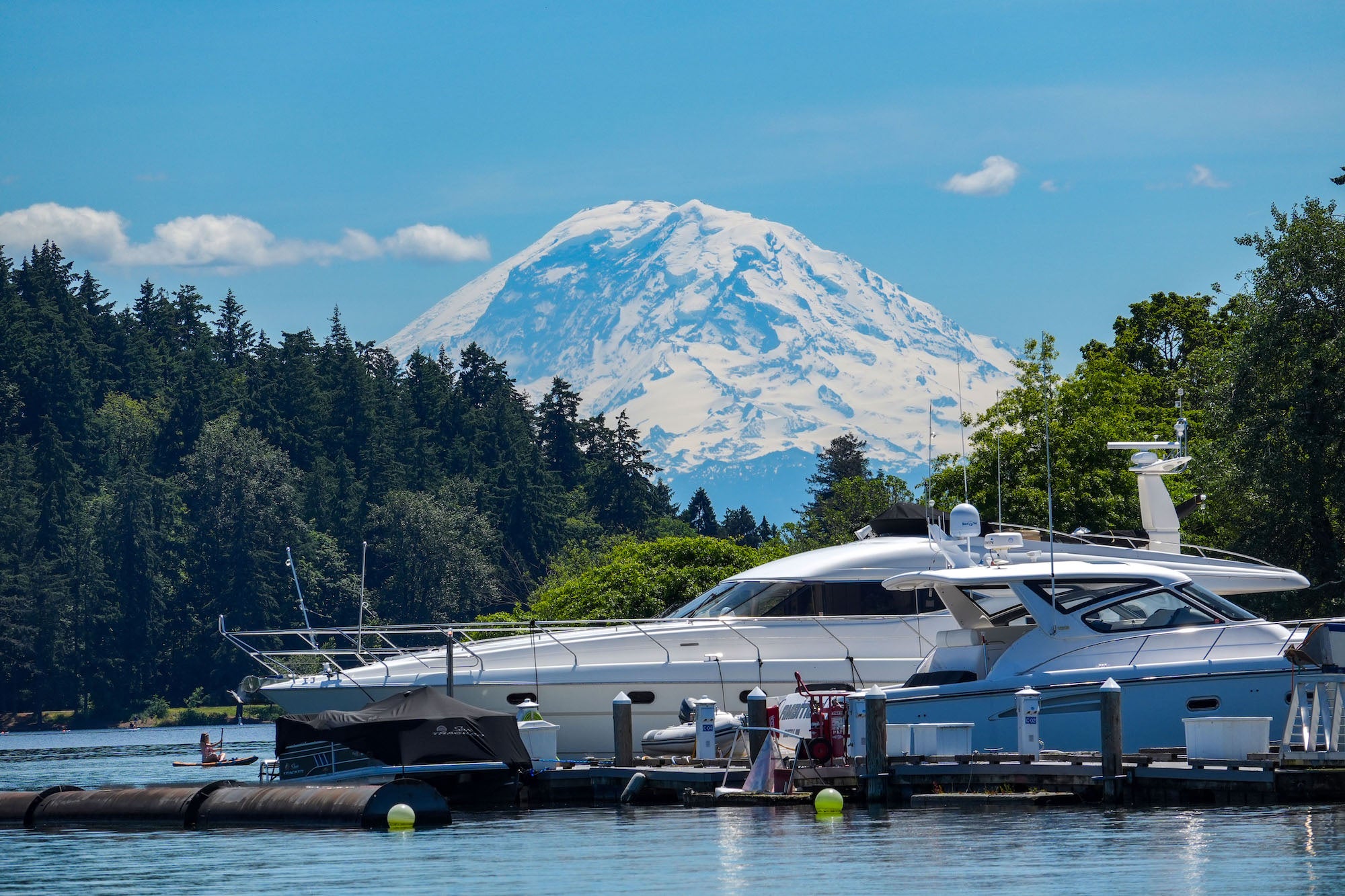 (Marina + Mt. Rainier) A sleek white yacht sits docked at a Pacific Northwest marina as the snow-covered summit of Mount Rainier rises dramatically above the surrounding evergreen forest &mdash; shot with the Sony RX10 V.