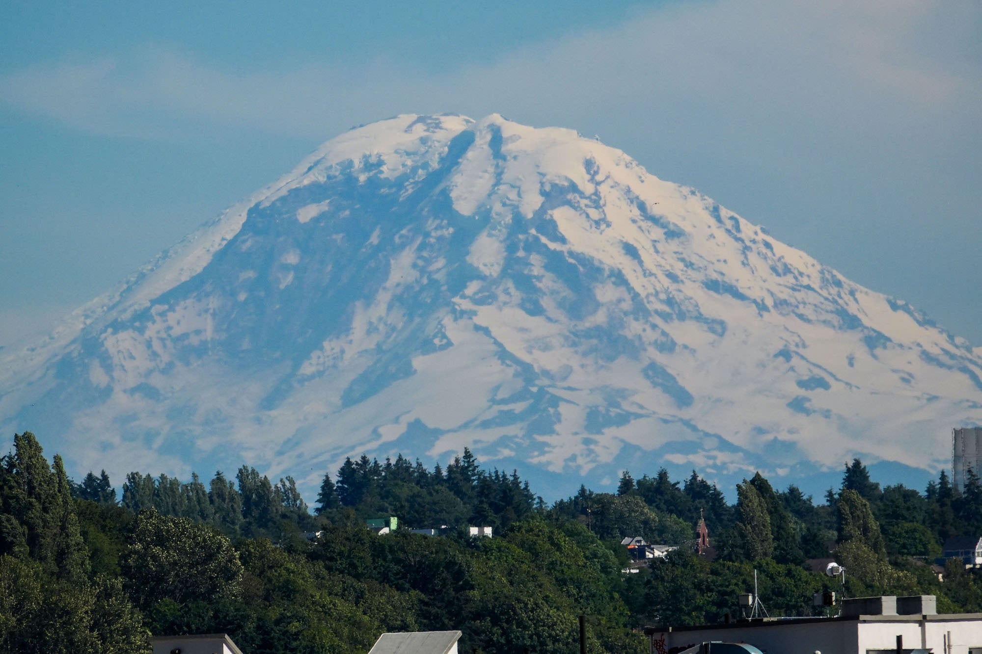 (Mt. Rainier over city) The snow-capped peak of Mount Rainier dominates the horizon above a Seattle neighborhood's rooftops and treetops, its glaciated summit filling the frame thanks to the RX10 V's powerful telephoto reach.
