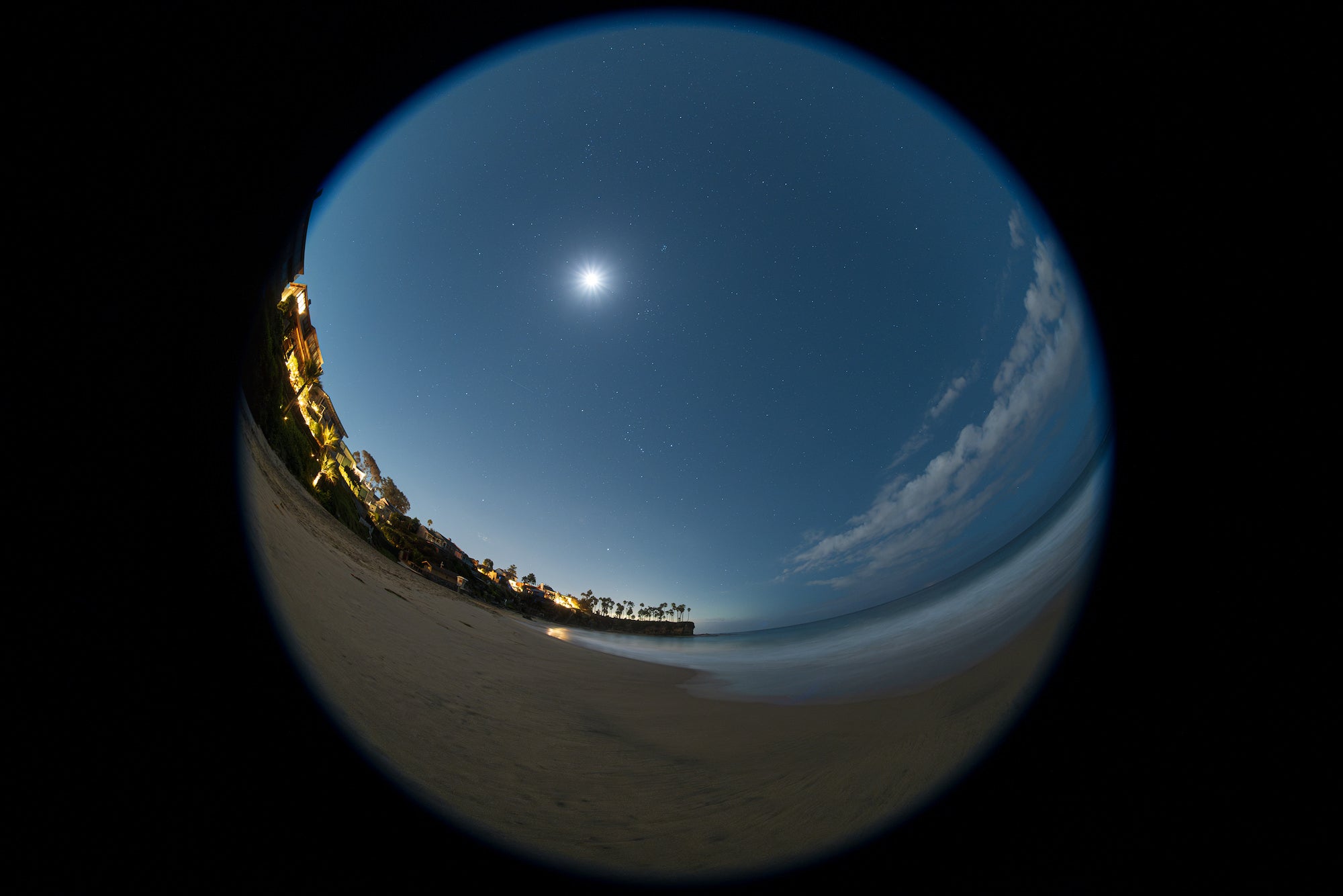 Circular fisheye view of a moonlit beach, ocean, and coastal homes under a starry sky, photographed by Stan Moniz.