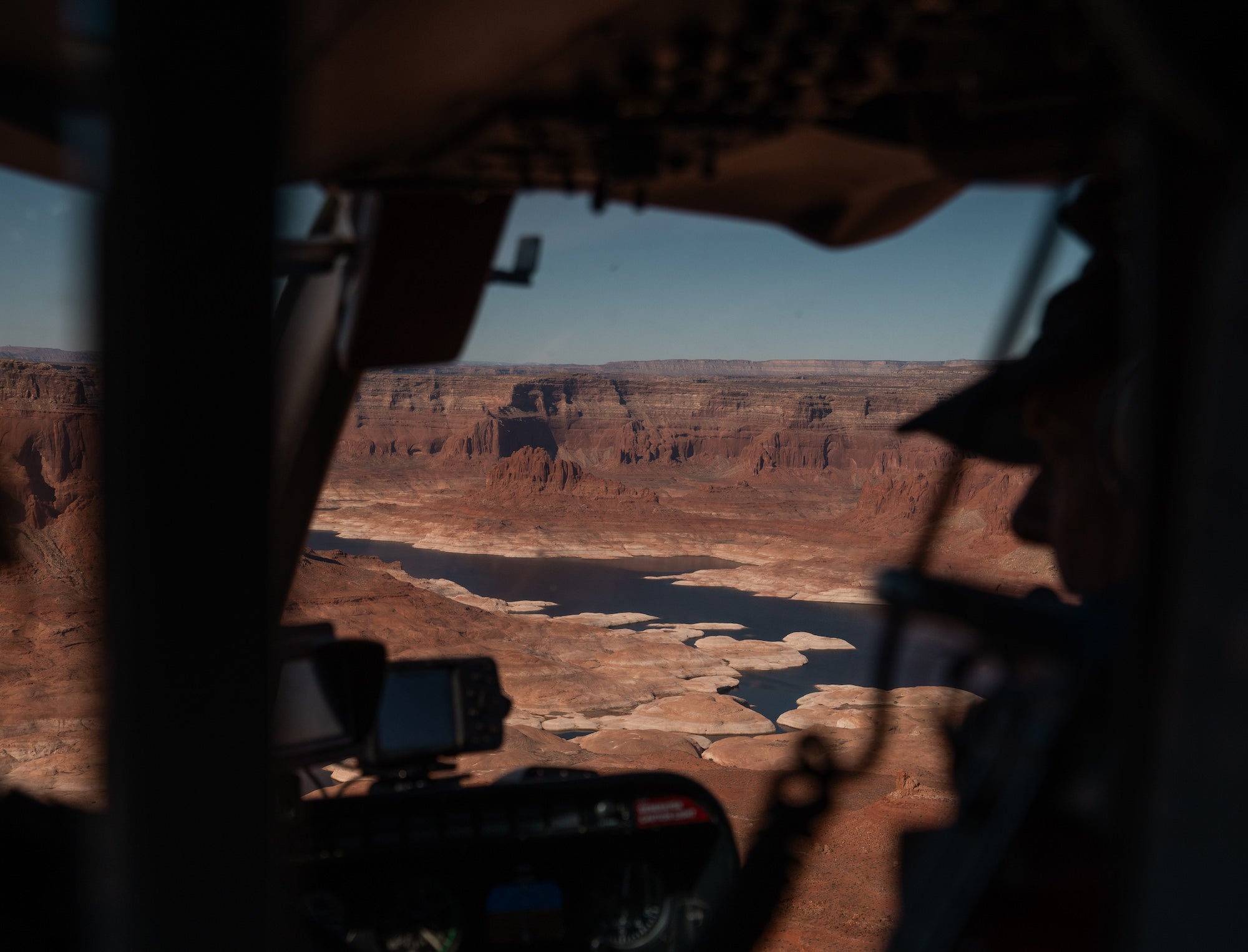 View from inside a helicopter of Lake Powell&rsquo;s dark water and pale exposed shoreline beneath red canyon cliffs. Photograph by Sydney Smolla.