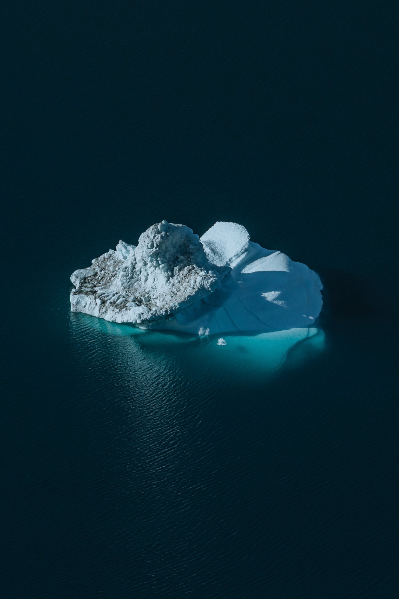 An aerial perspective captures a solitary iceberg floating in near-black deep water, its bright white and glacial-blue surfaces casting dramatic shadows. Photographed by Eyr&uacute;n Lyd&iacute;a S&aelig;varsd&oacute;ttir with the Sony FE 100-400mm f/4.5-5.6 G Master.