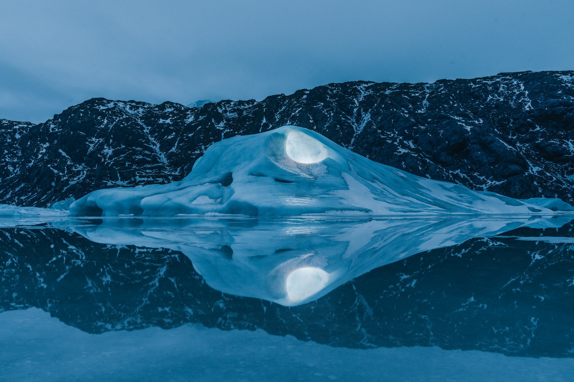 A glowing iceberg reflects perfectly on the mirror-still surface of a glacial lake at twilight, framed by dark, snow-dusted mountains. Photographed by Eyr&uacute;n Lyd&iacute;a S&aelig;varsd&oacute;ttir with the Sony FE 16-35mm f/2.8 G Master.