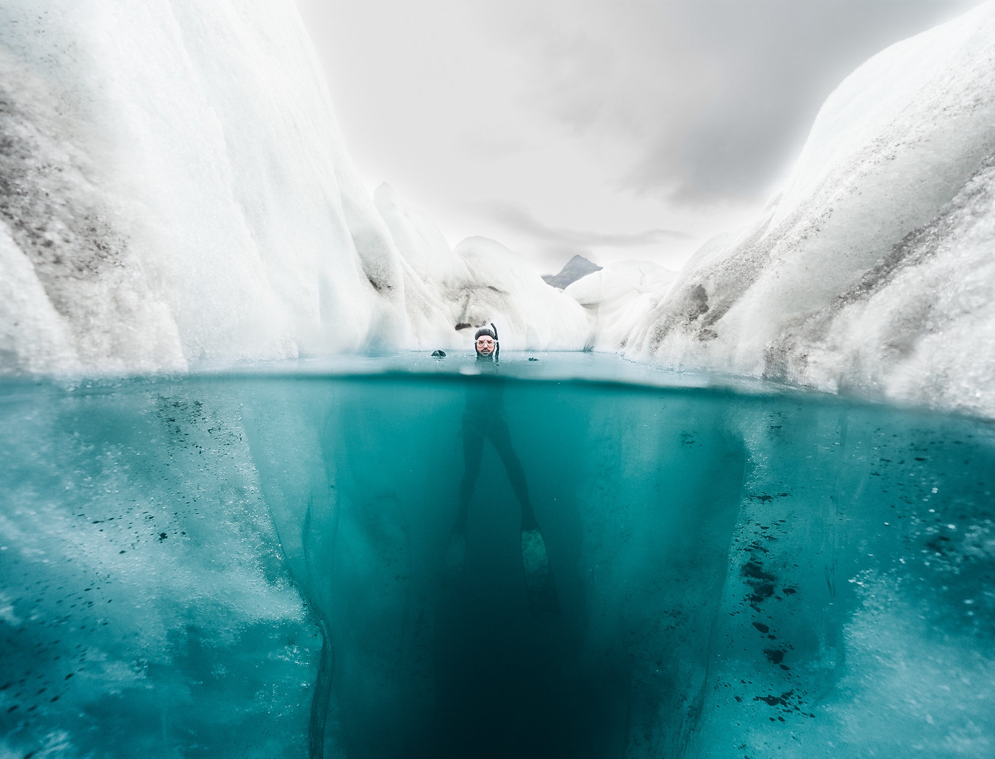 A split above/below-water view reveals a snorkeler suspended between towering walls of ice in brilliant glacial-blue meltwater. Photographed by Eyr&uacute;n Lyd&iacute;a S&aelig;varsd&oacute;ttir with the Sony FE 16-35mm f/2.8 G Master.