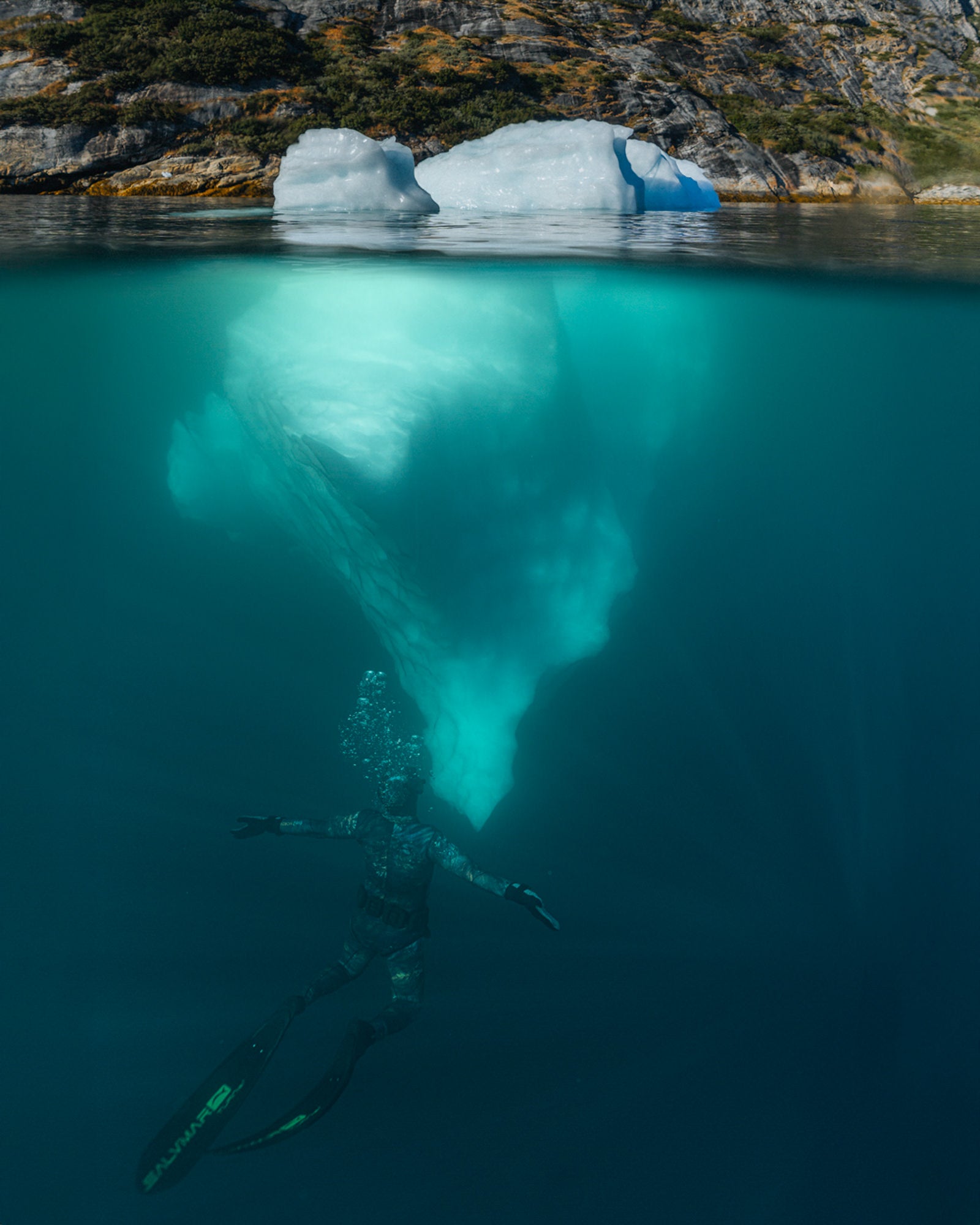 A split-level underwater image shows a freediver descending beneath a small iceberg, revealing the glowing submerged mass against rocky Greenlandic shoreline above. Photographed by Eyr&uacute;n Lyd&iacute;a S&aelig;varsd&oacute;ttir with the Sony FE 16-35mm f/2.8 G Master.