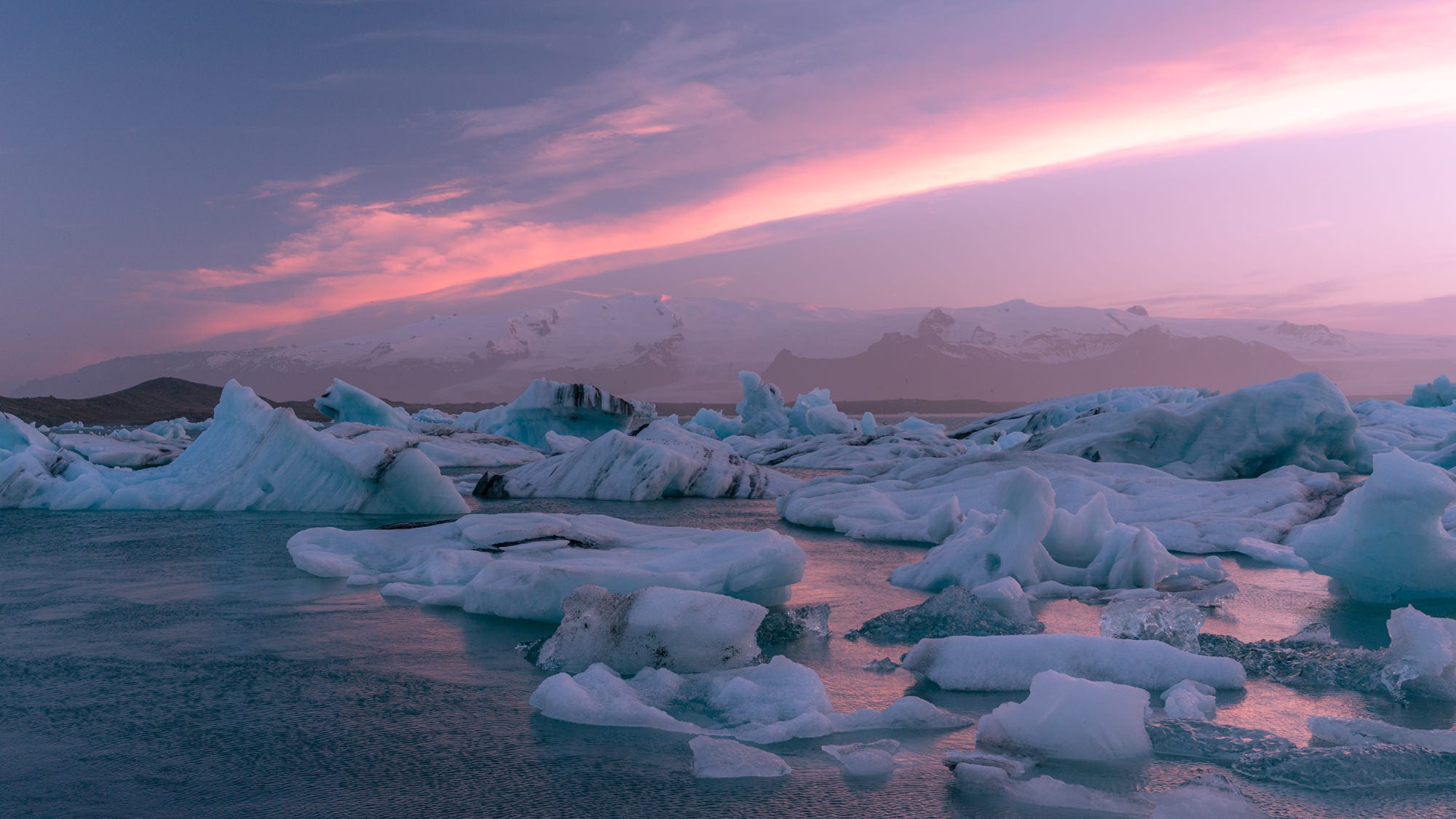 Dozens of icebergs drift across a glacial lagoon beneath a breathtaking pink and purple sunset sky, with snow-capped mountains glowing on the horizon. Photographed by Eyr&uacute;n Lyd&iacute;a S&aelig;varsd&oacute;ttir with the Sony FE 24-70mm f/2.8 G Master II.