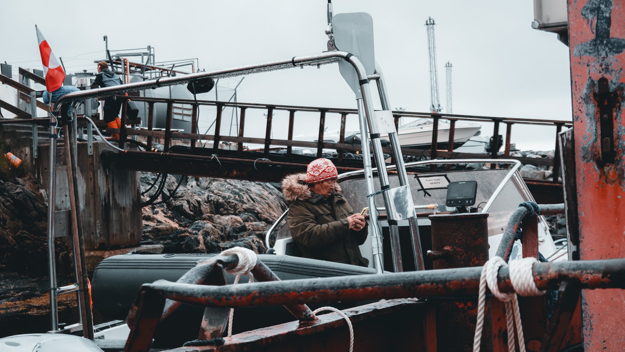 A local woman in a red patterned hat checks her phone aboard a small boat at a weathered Arctic harbor dock, capturing everyday life in Greenland. Photographed by Eyr&uacute;n Lyd&iacute;a S&aelig;varsd&oacute;ttir with the Sony RX100 VII.