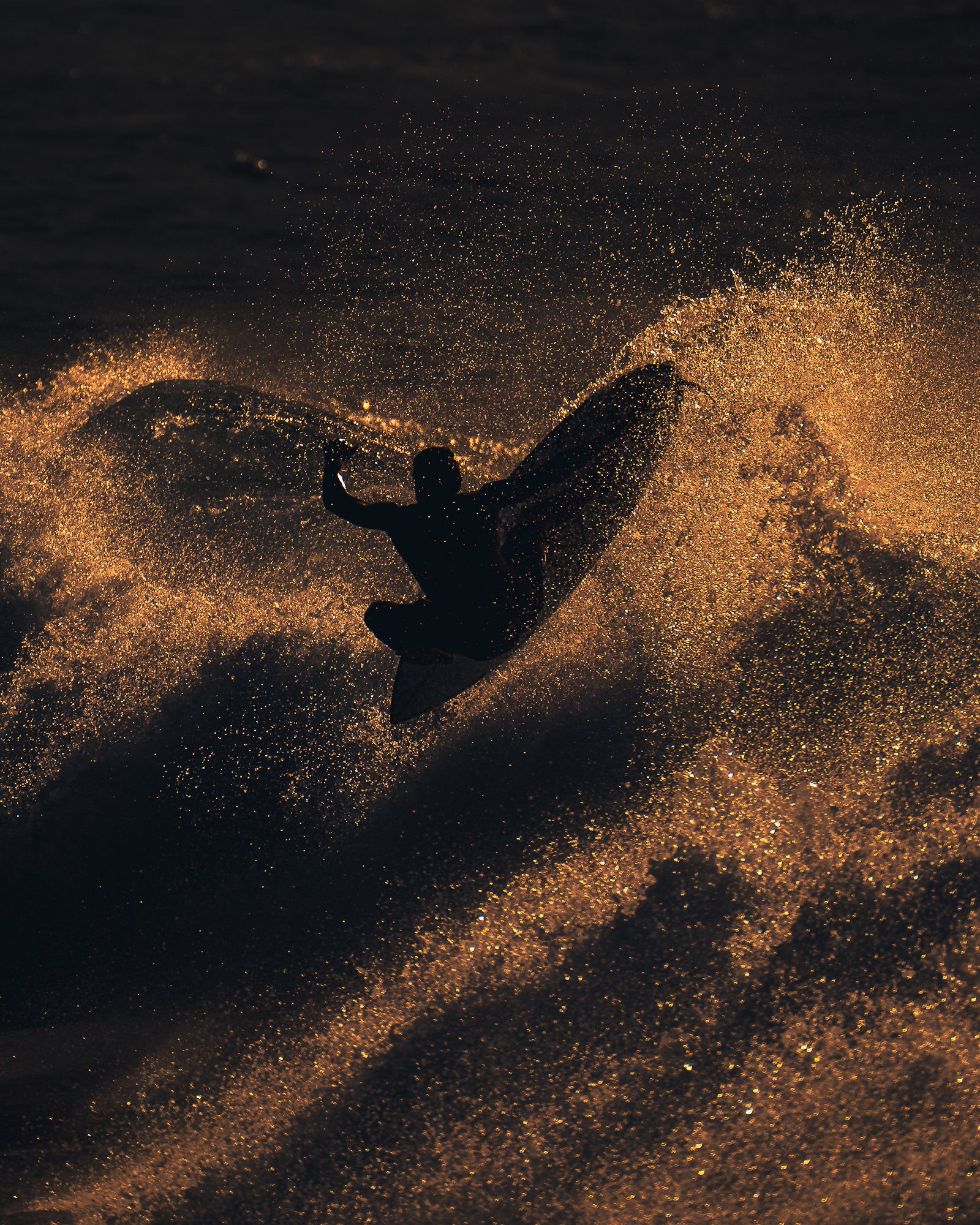 A surfer launches an aerial maneuver surrounded by an explosion of golden backlit spray, the board and body silhouetted against glowing mist.