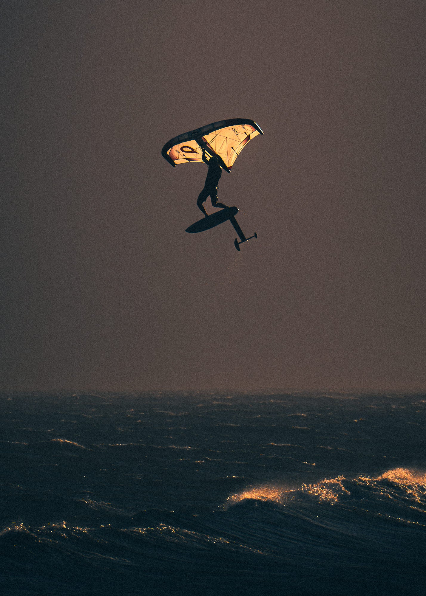 A wing foiler soars high above dark ocean swells, their inflated wing and hydrofoil board clearly silhouetted against a moody dusky sky.