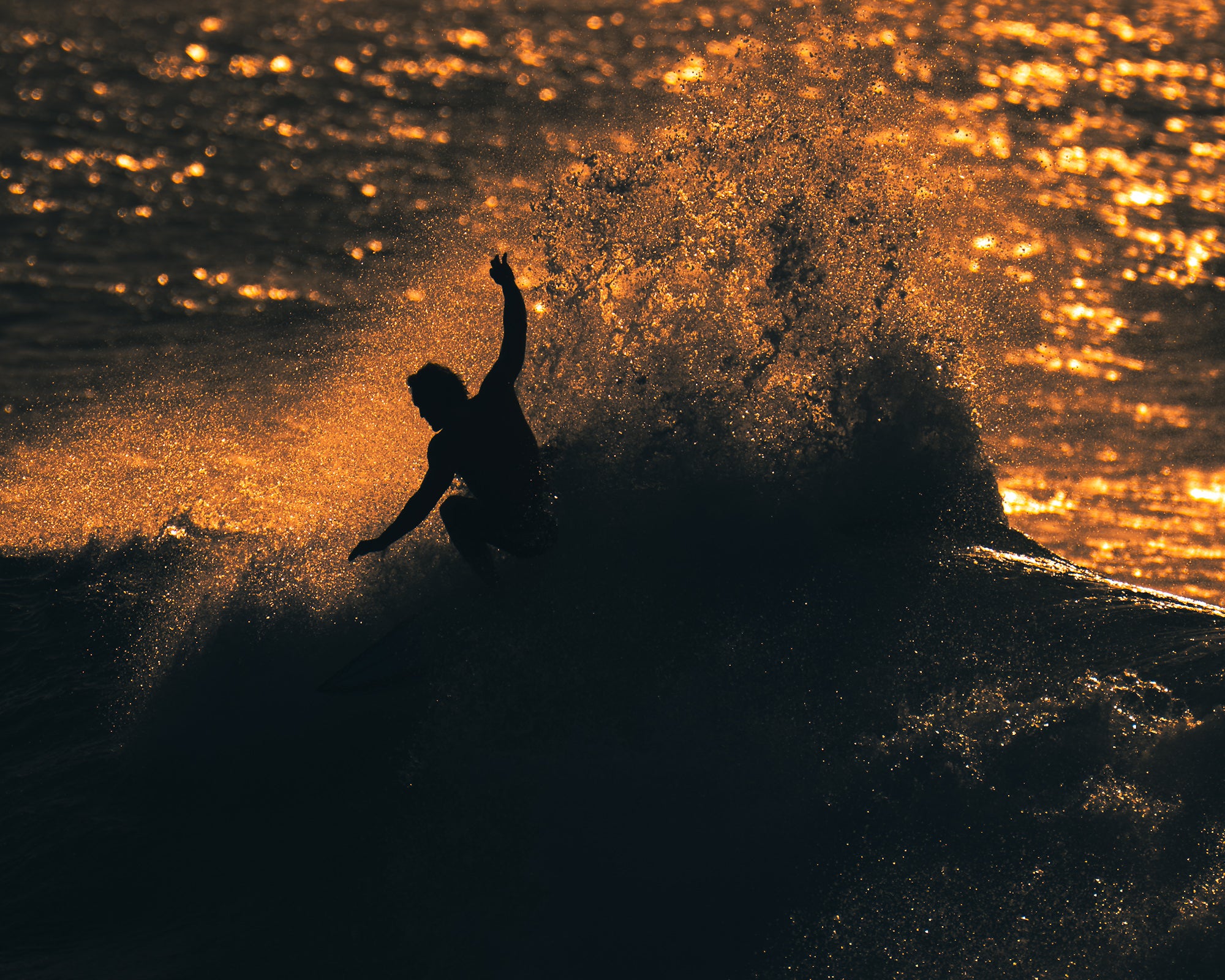 A surfer mid-maneuver raises one arm as a massive curtain of golden spray erupts behind them, the ocean surface shimmering with warm light.