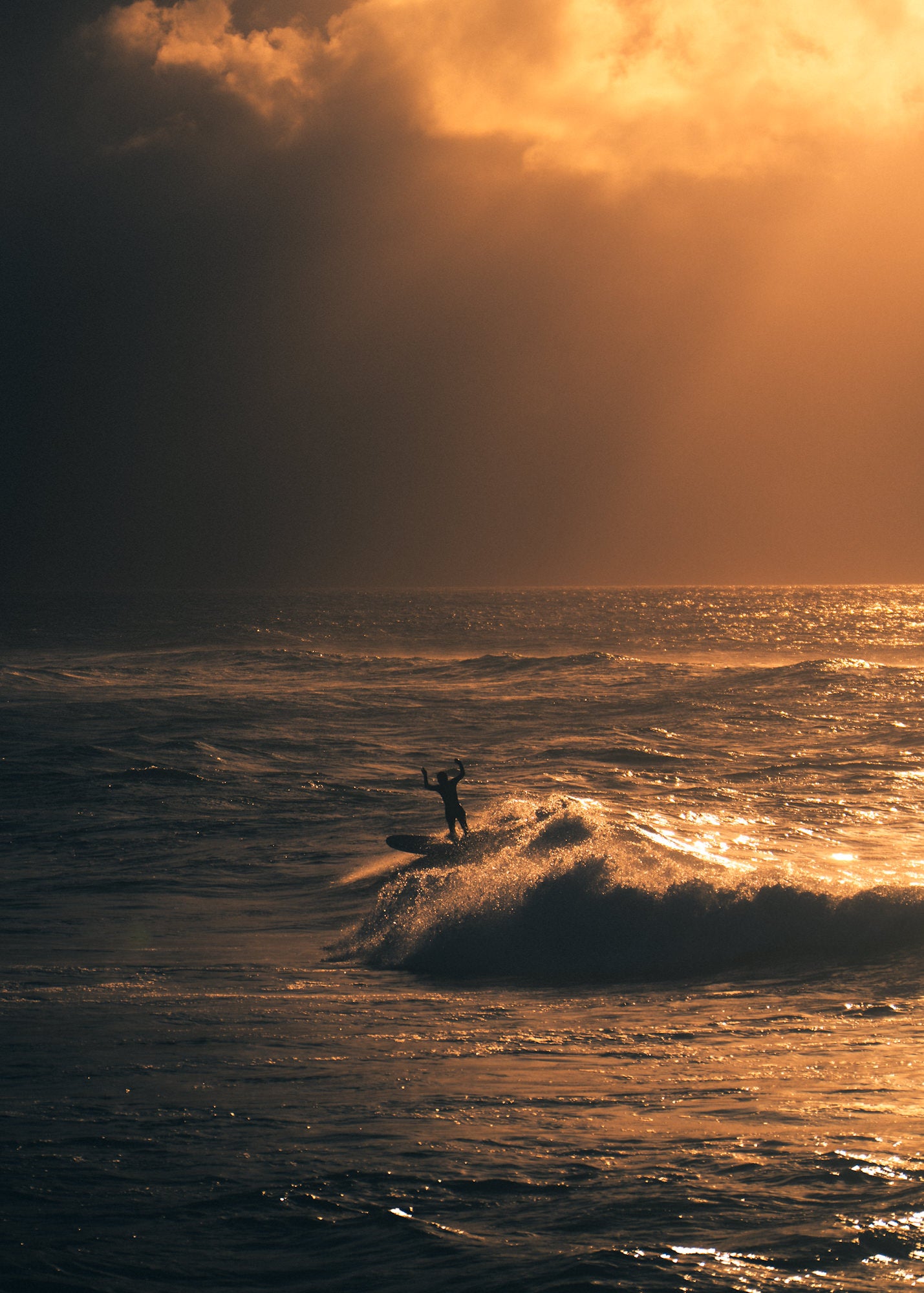 A surfer raises both arms in celebration while riding a glowing wave beneath a stormy golden sky, perfectly isolated against the open ocean.