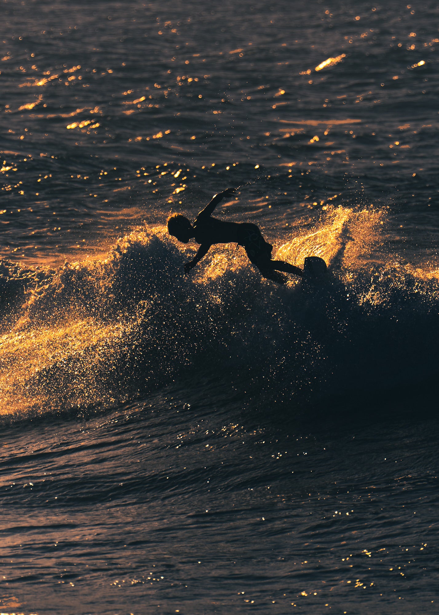 A surfer wipes out in spectacular fashion at golden hour, their silhouette airborne above the board as sun-kissed spray explodes around them.