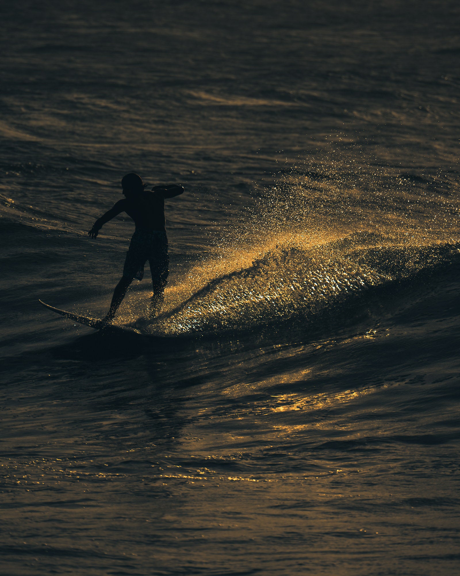 A surfer carves a smooth bottom turn at dusk, trailing a sweeping arc of illuminated golden spray across a darkened ocean surface.