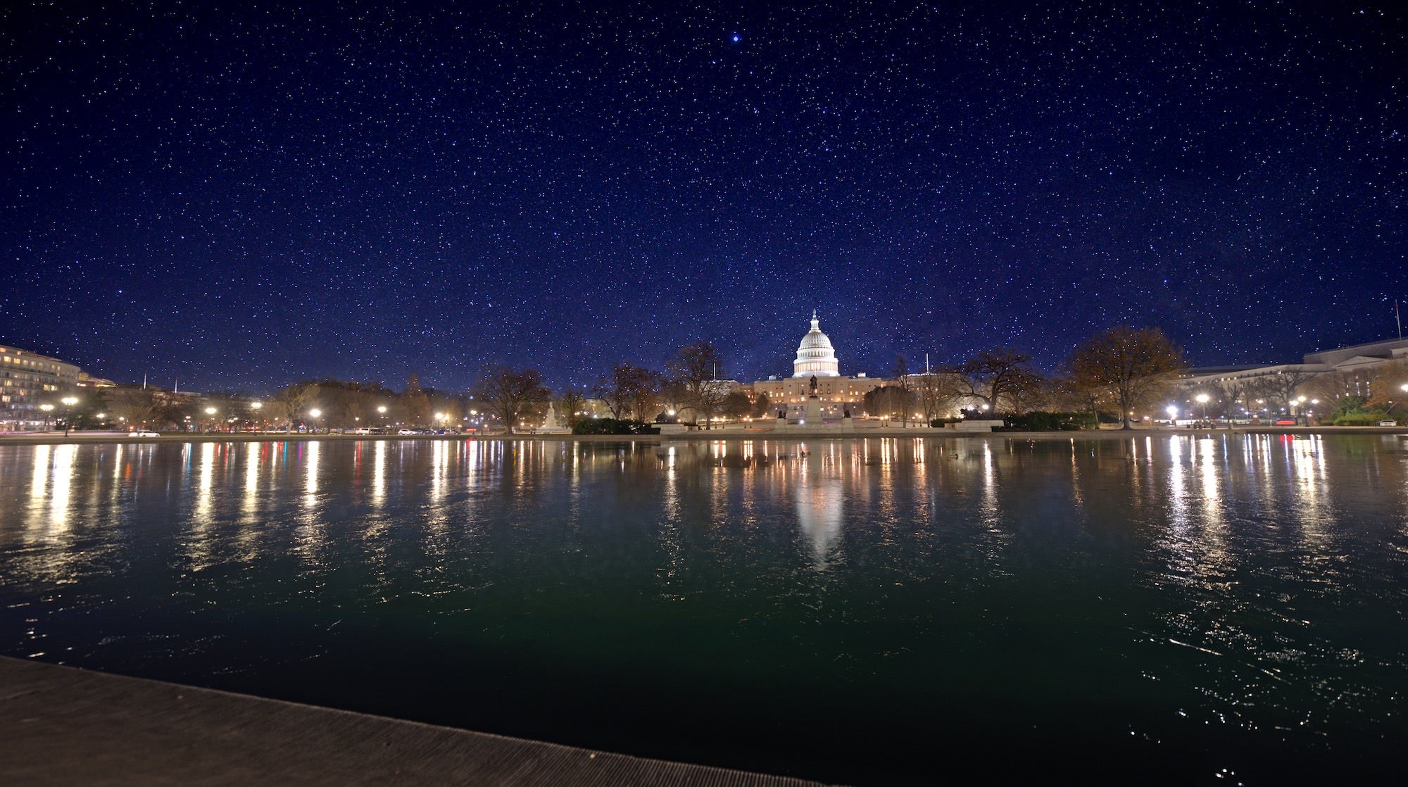 U.S. Capitol across the Reflecting Pool at night, dome lit and stars overhead with city lights mirrored in the water.