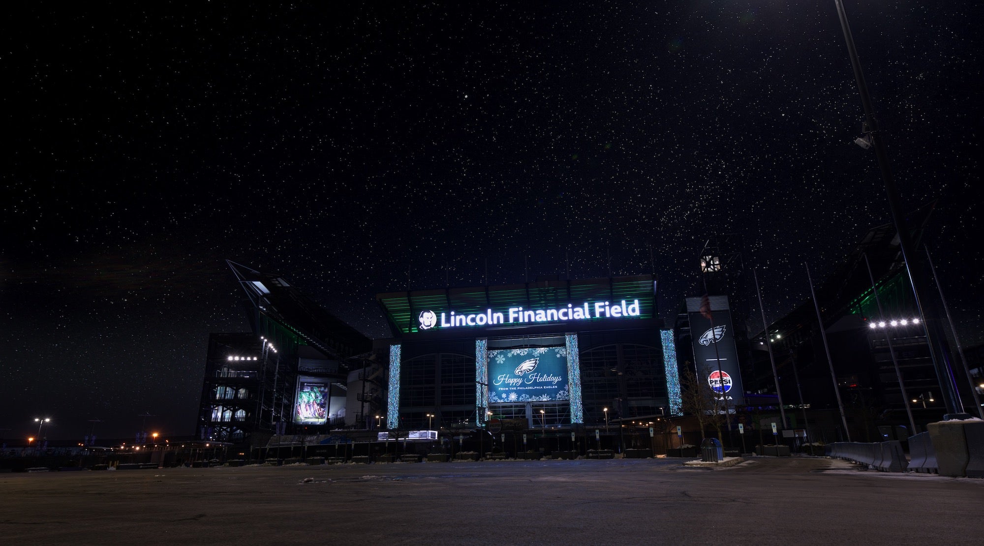 Lincoln Financial Field illuminated at night under a starry sky, holiday graphics on the facade and an empty plaza in front.