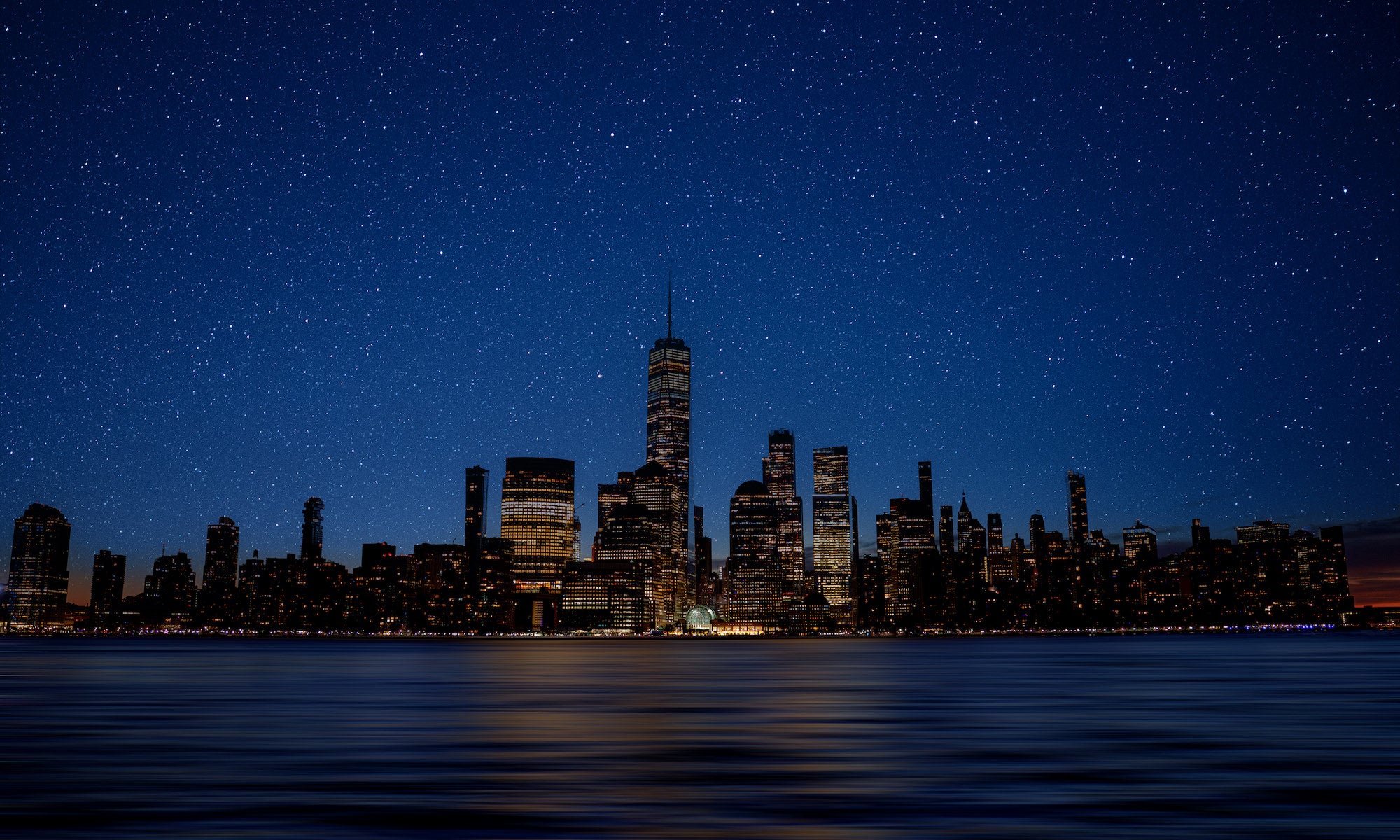 Panoramic Lower Manhattan at night, One World Trade Center centered beneath a starry sky with river reflections.