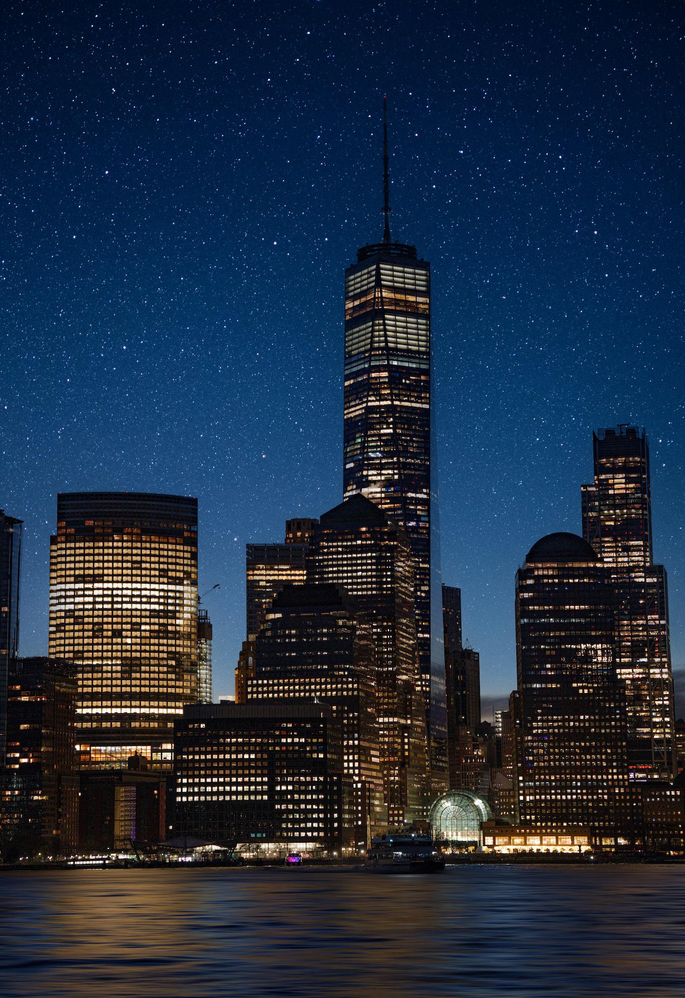 Lower Manhattan skyline with One World Trade Center under a star-filled night sky, windows glowing above the Hudson River.