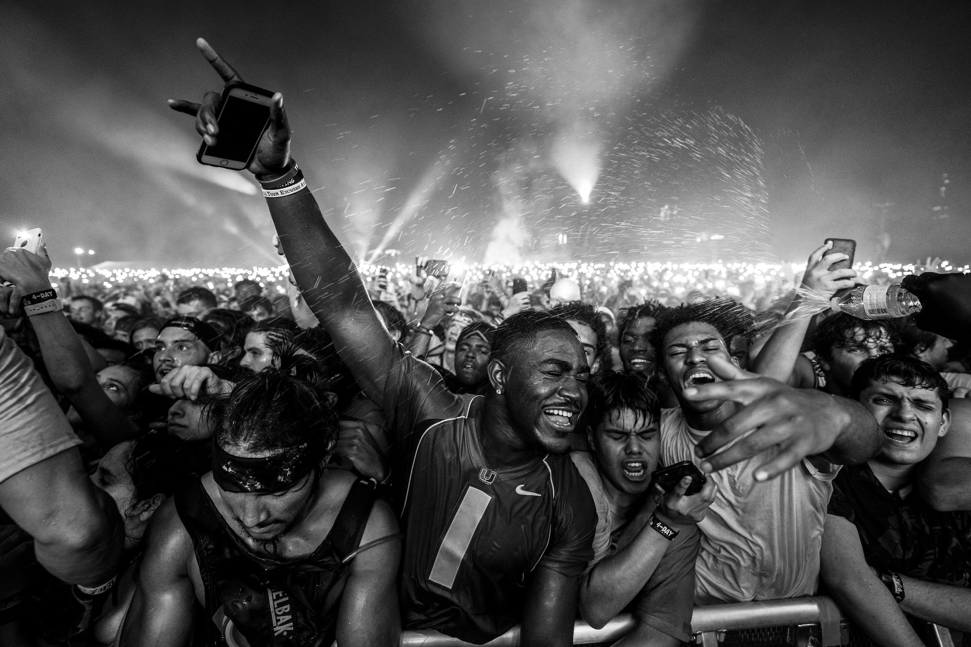 Packed concert crowd cheering near barricade as water sprays and lights glow.