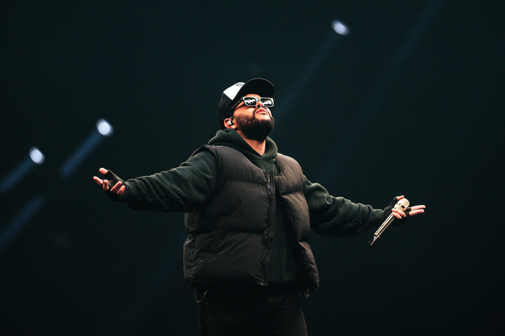 Performer in dark vest and cap stands arms wide on a dim stage, holding a mic, spotlights cutting through the dark.