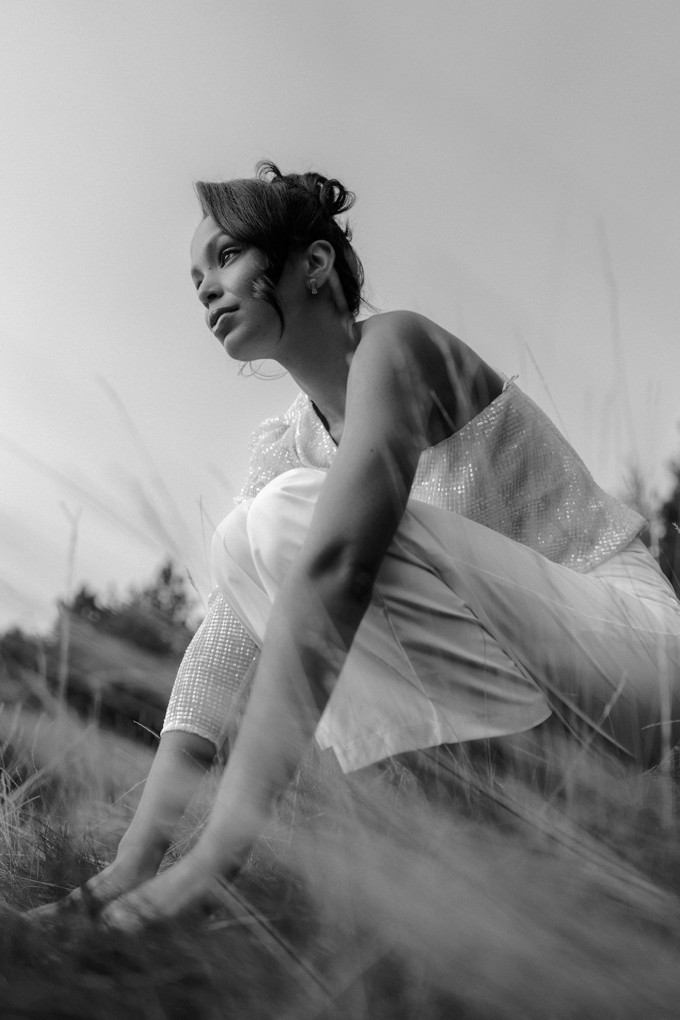 Low-angle black-and-white portrait; person crouching in tall grass, thoughtful gaze.