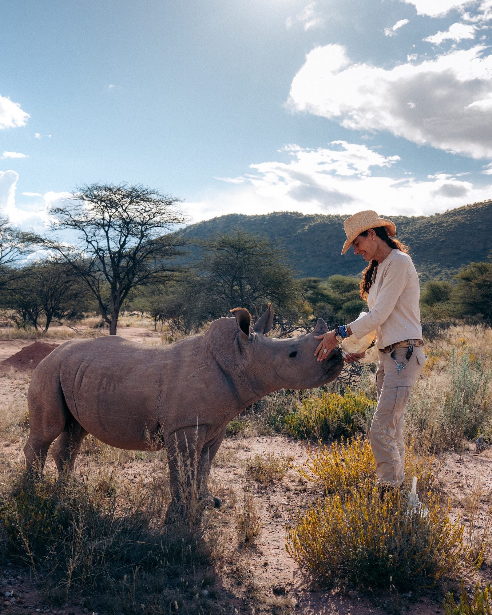 A person in a straw hat gently touches a rhino&rsquo;s face in a dry grassland, with low wooded hills behind. Photo by Jay Baumann. Alpha 7 IV. 24-70mm f/2.8 G Master II. 1/2000-sec. f/5.6. ISO 800.