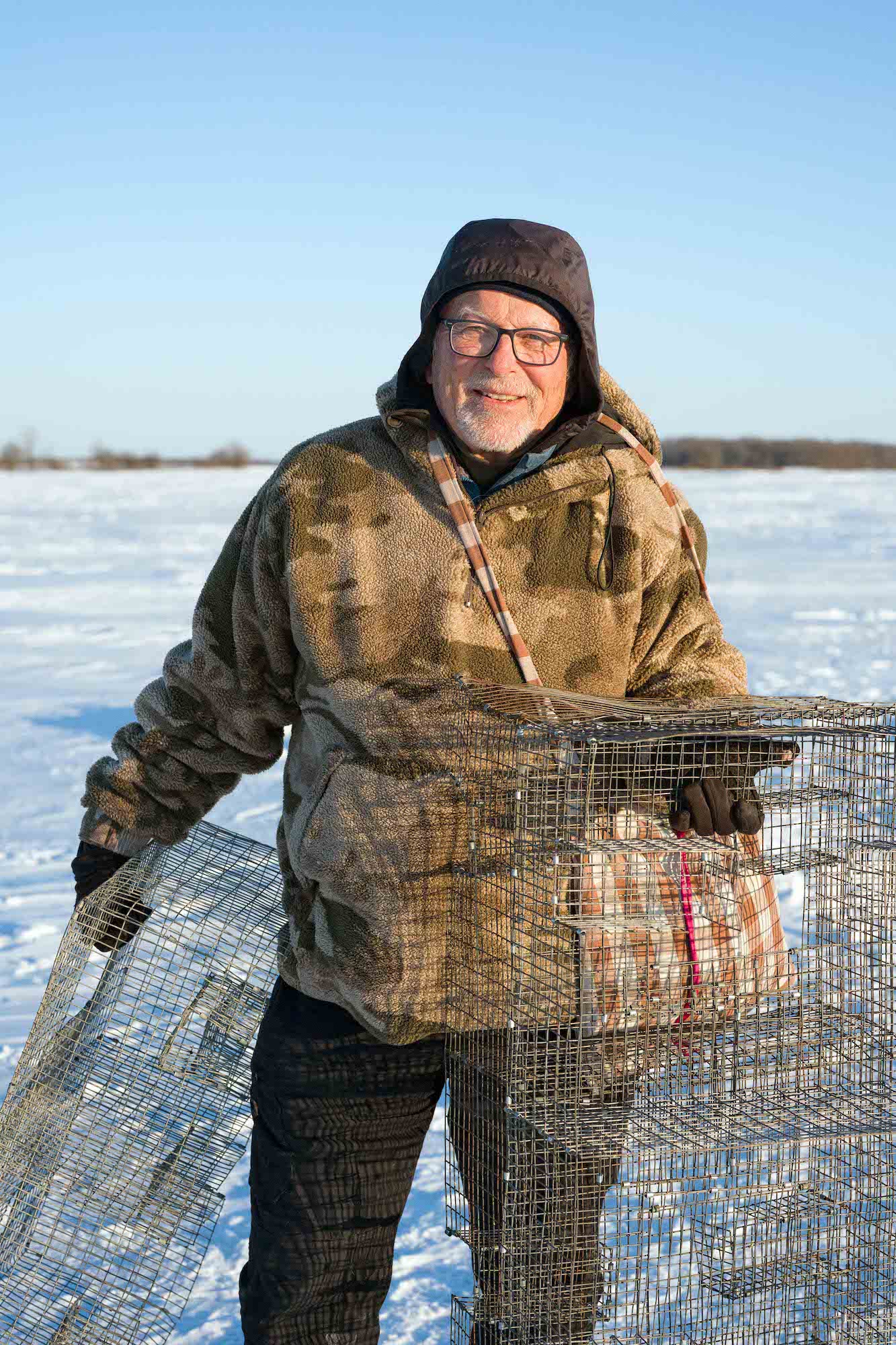 Person in heavy winter clothing smiling while holding wire cages on a snowy field.