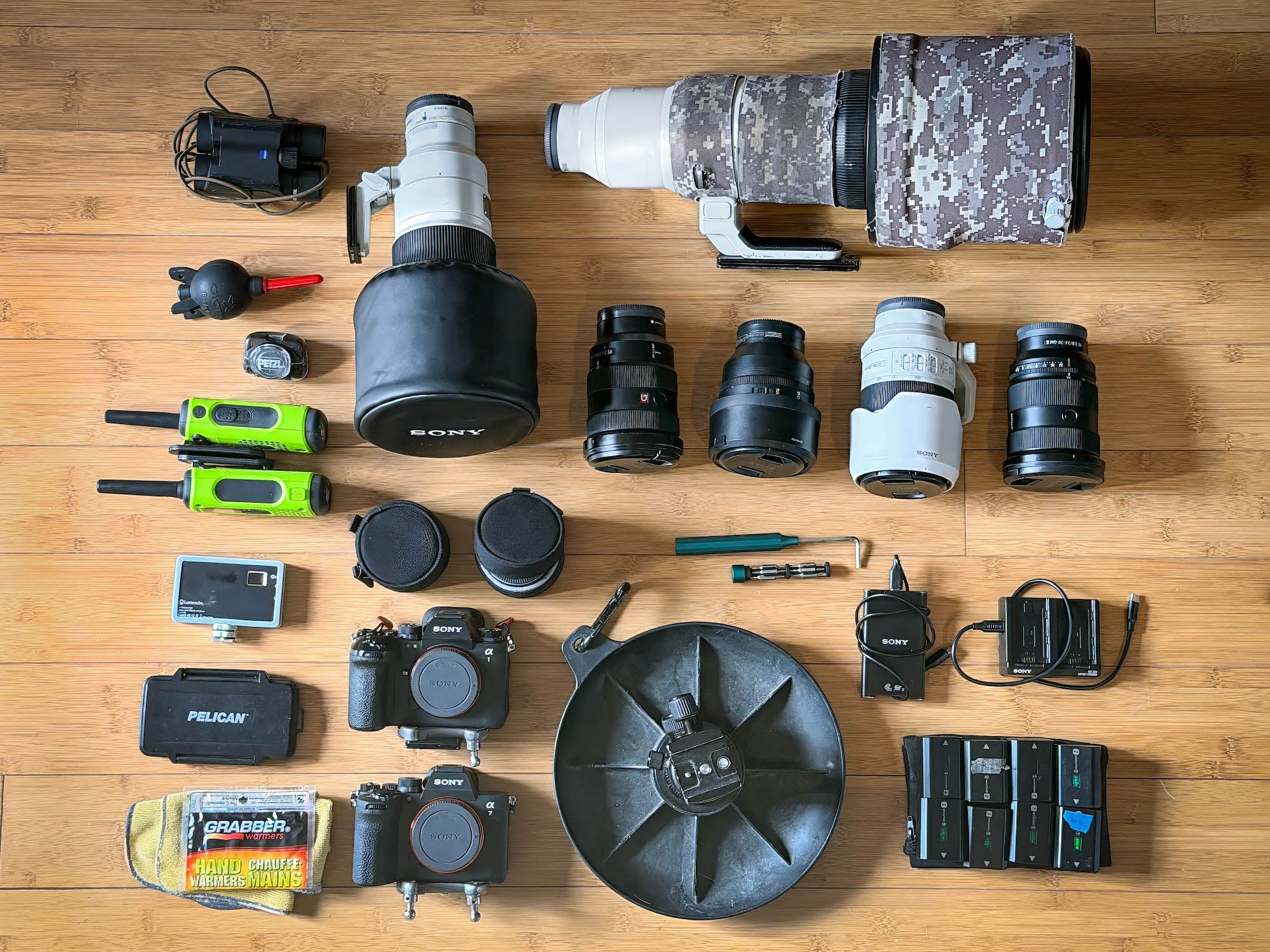 Flat lay of wildlife photo gear&mdash;cameras, long lenses, radios, batteries&mdash;on wood floor.