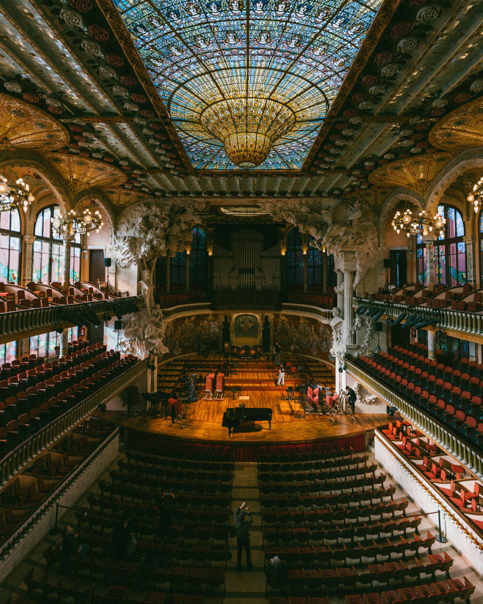 An ornate concert hall viewed from an upper balcony, with tiered rows of red seats, a grand stained-glass ceiling and chandelier, sculptural columns, and a warmly lit stage holding a grand piano and music stands. Photograph by Ming Han.