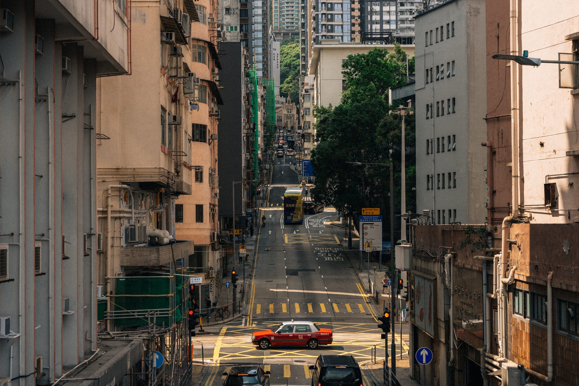 A steep urban street runs between closely packed mid-rise buildings, with a red taxi crossing a yellow-marked intersection and a double-decker bus climbing the hill in the distance. Photograph by Ming Han.
