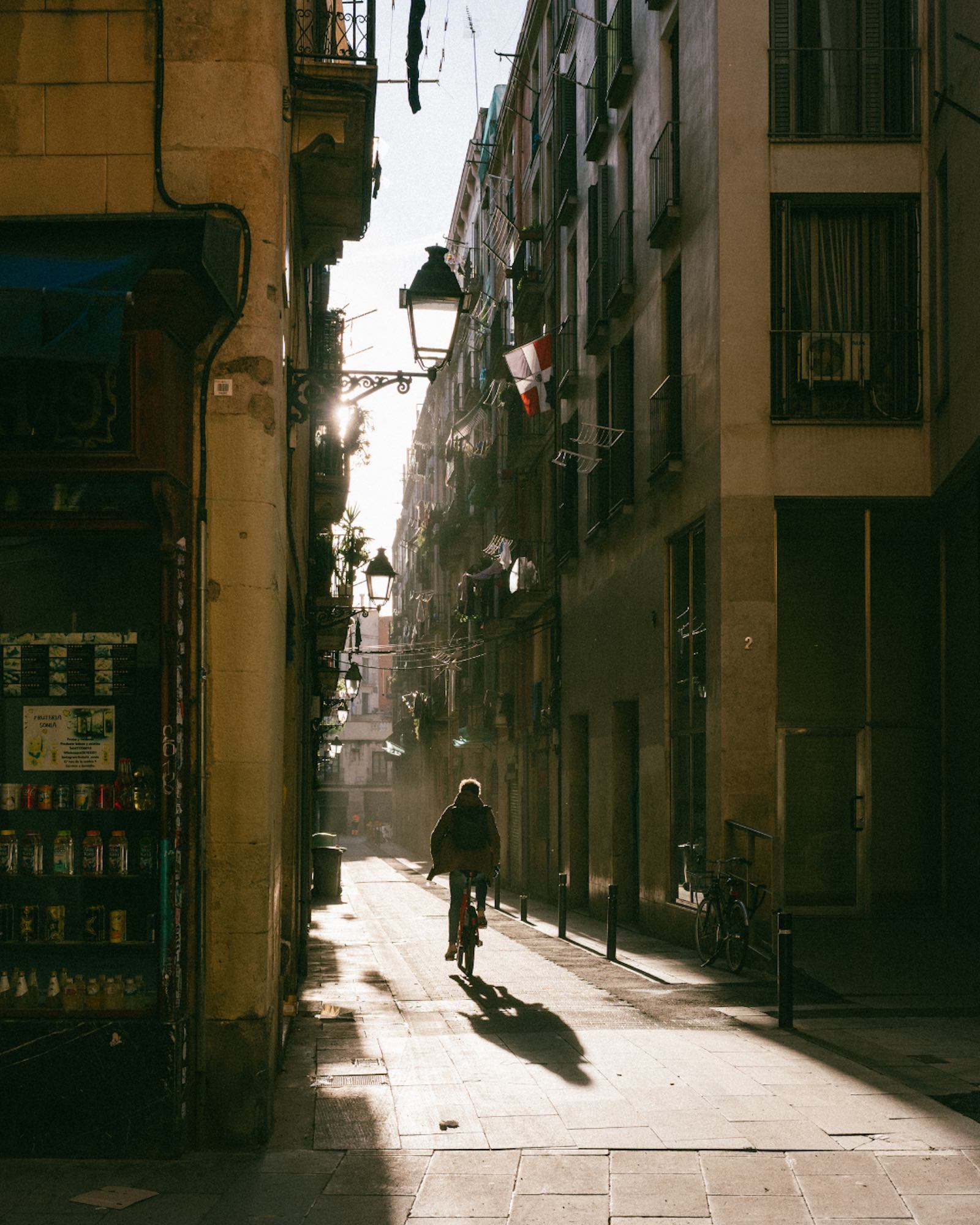 A cyclist rides through a narrow, sunlit street lined with tall apartment buildings, balconies, hanging laundry, and old-style streetlamps. Long shadows stretch across the pavement. Photograph by Ming Han.