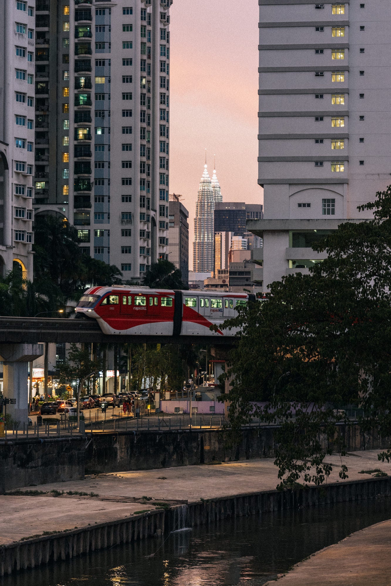 A red-and-white elevated train passes between tall residential towers at dusk, with a canal in the foreground and a brightly lit, twin-spired skyscraper centered in the distance. Photograph by Ming Han.