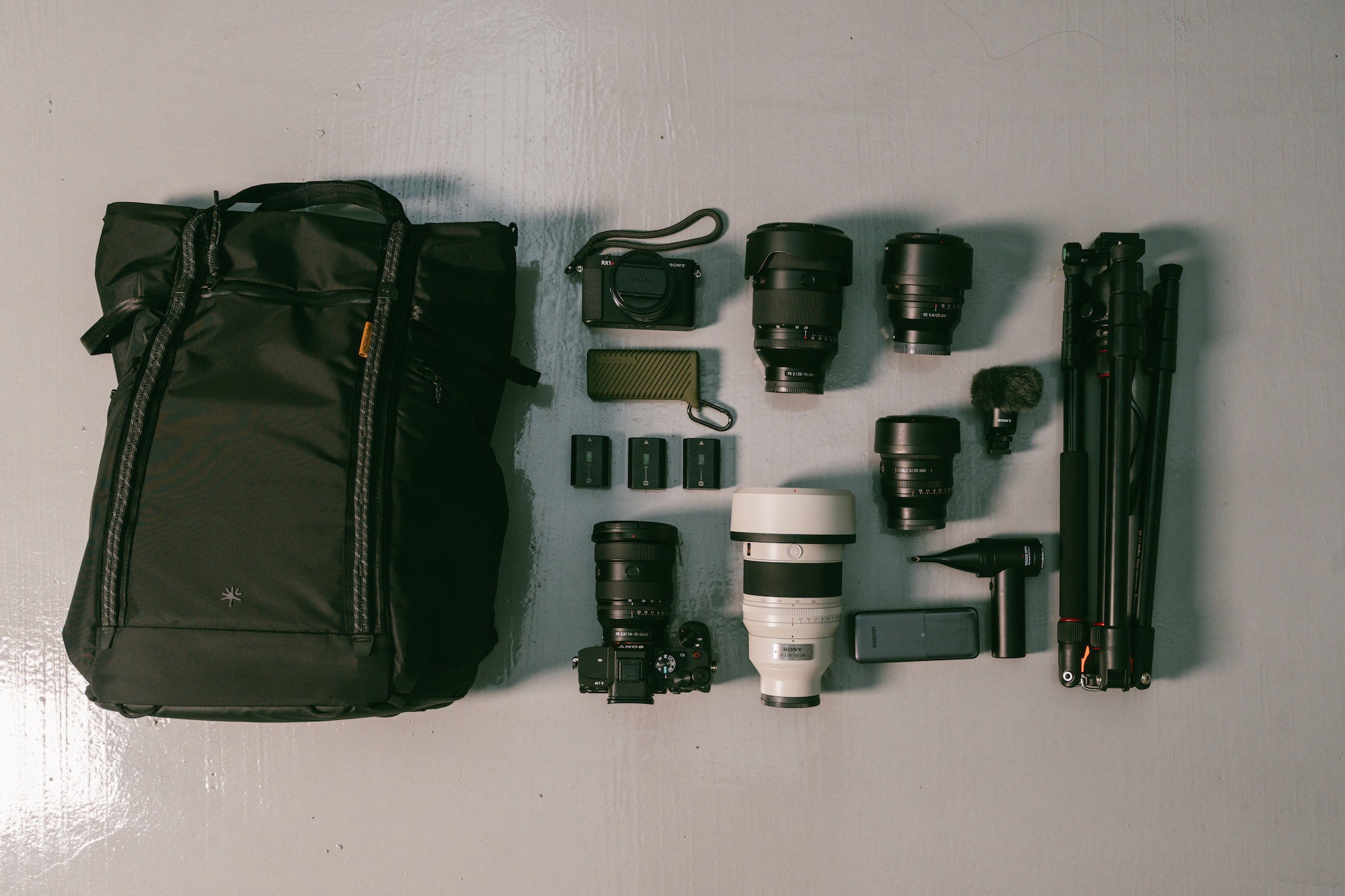 Overhead view of Ming Hang&rsquo;s black camera backpack laid out beside a compact camera, several interchangeable lenses, three batteries, a camera body, a large white telephoto lens, accessories, and a folded tripod. Photograph by Ming Hang.