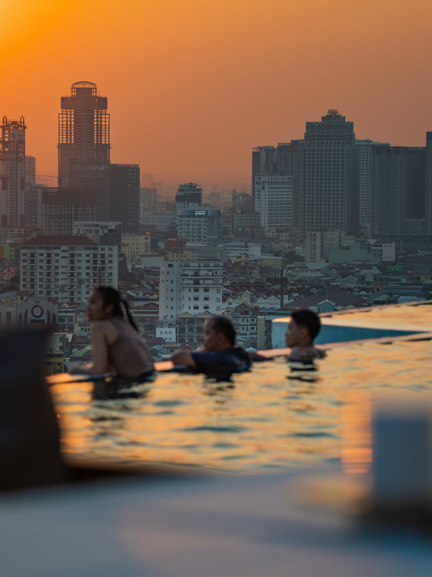 Three people relax in a rooftop infinity pool at sunset, with a dense city skyline in the background. Photograph by Nat Dabek.