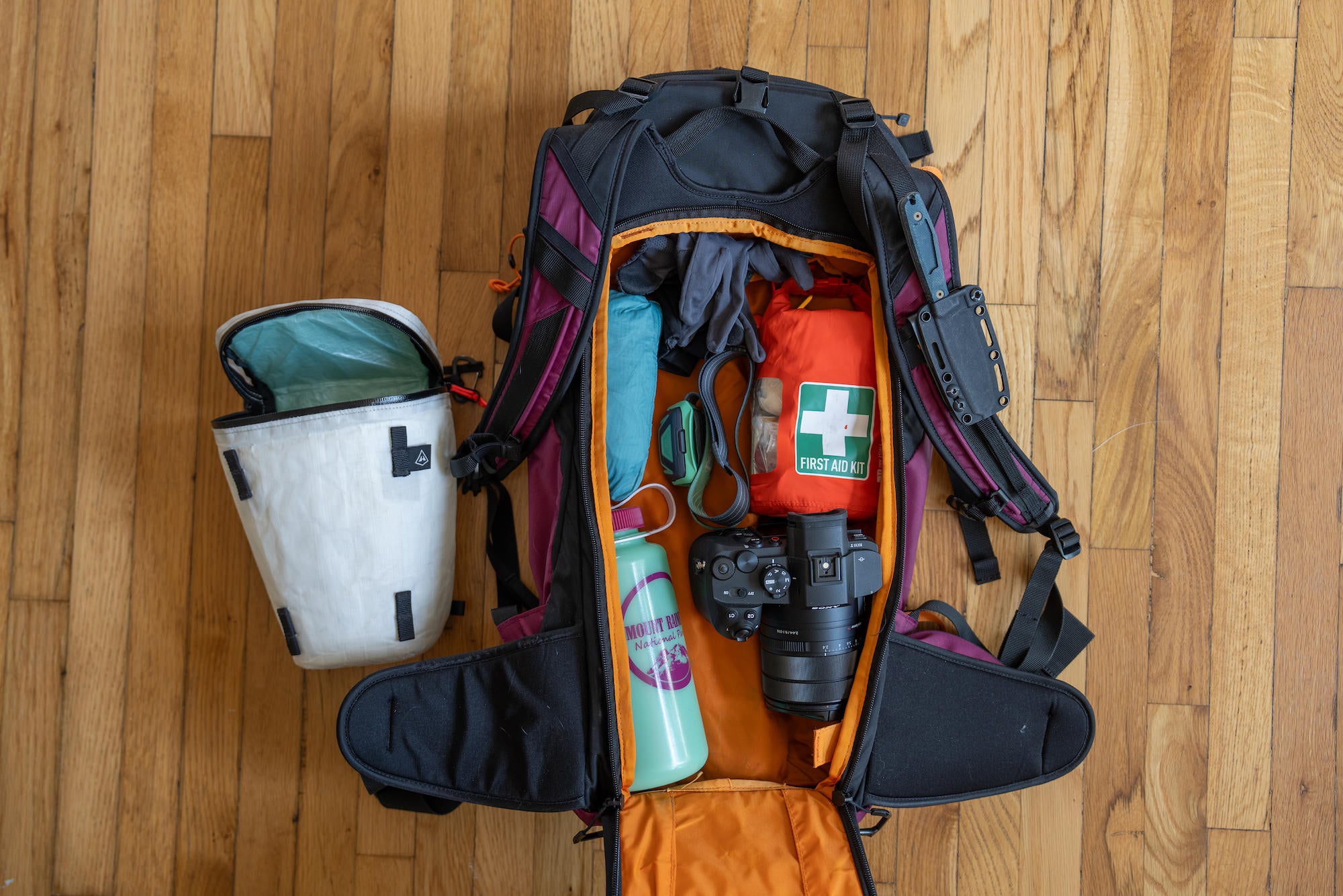 A fully packed camera backpack lays open to reveal a Sony camera, first aid kit, water bottle, and compact accessories &mdash; Nate Luebbe's complete Sony RX10 V field kit.