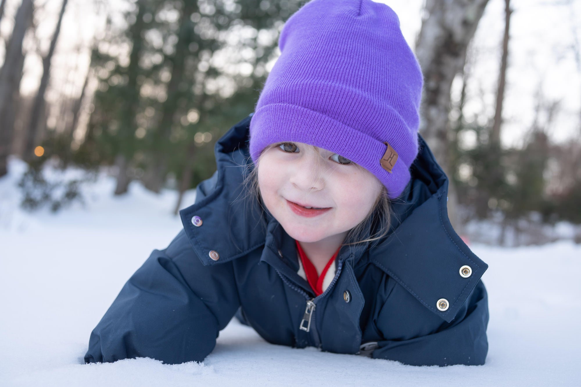 Child in a purple beanie and winter coat lying in the snow, smiling.