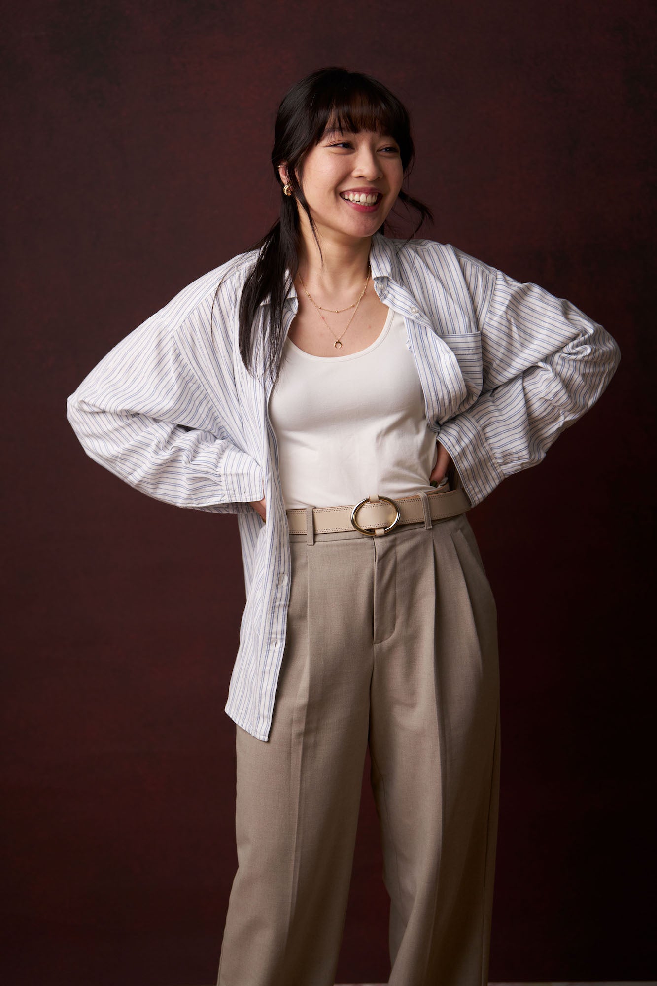 Full-length portrait of a woman laughing with hands on hips, striped oversized shirt and beige trousers, warm studio background.