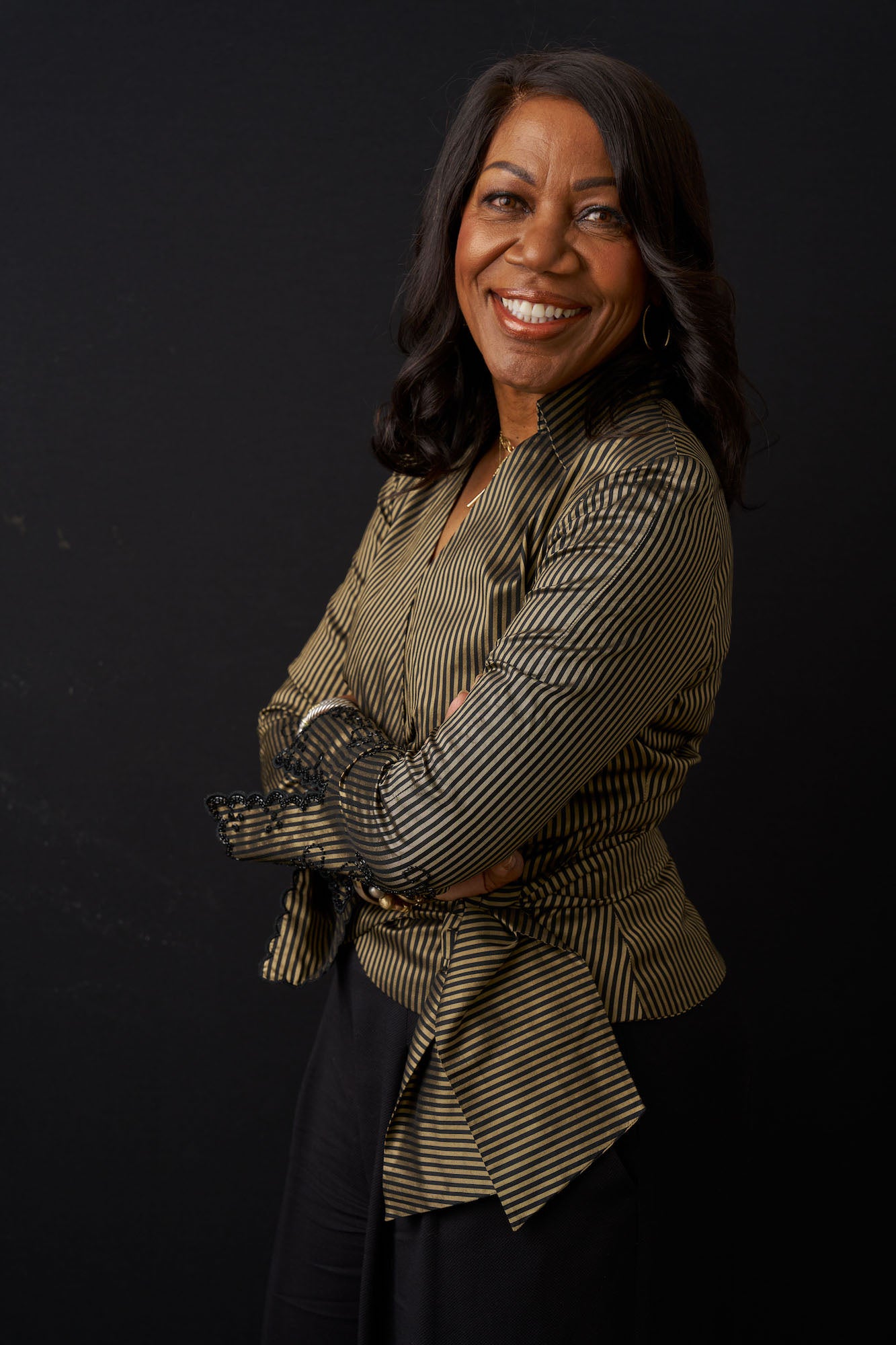 Studio portrait of a smiling woman in a striped blouse with arms crossed, black background.