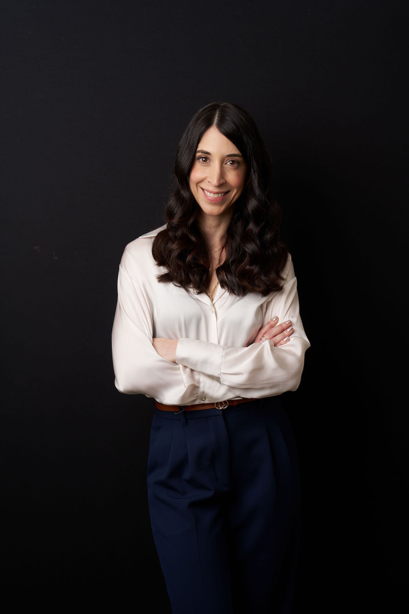 Three-quarter studio portrait of a woman in a cream blouse and navy trousers, arms crossed against a black backdrop.