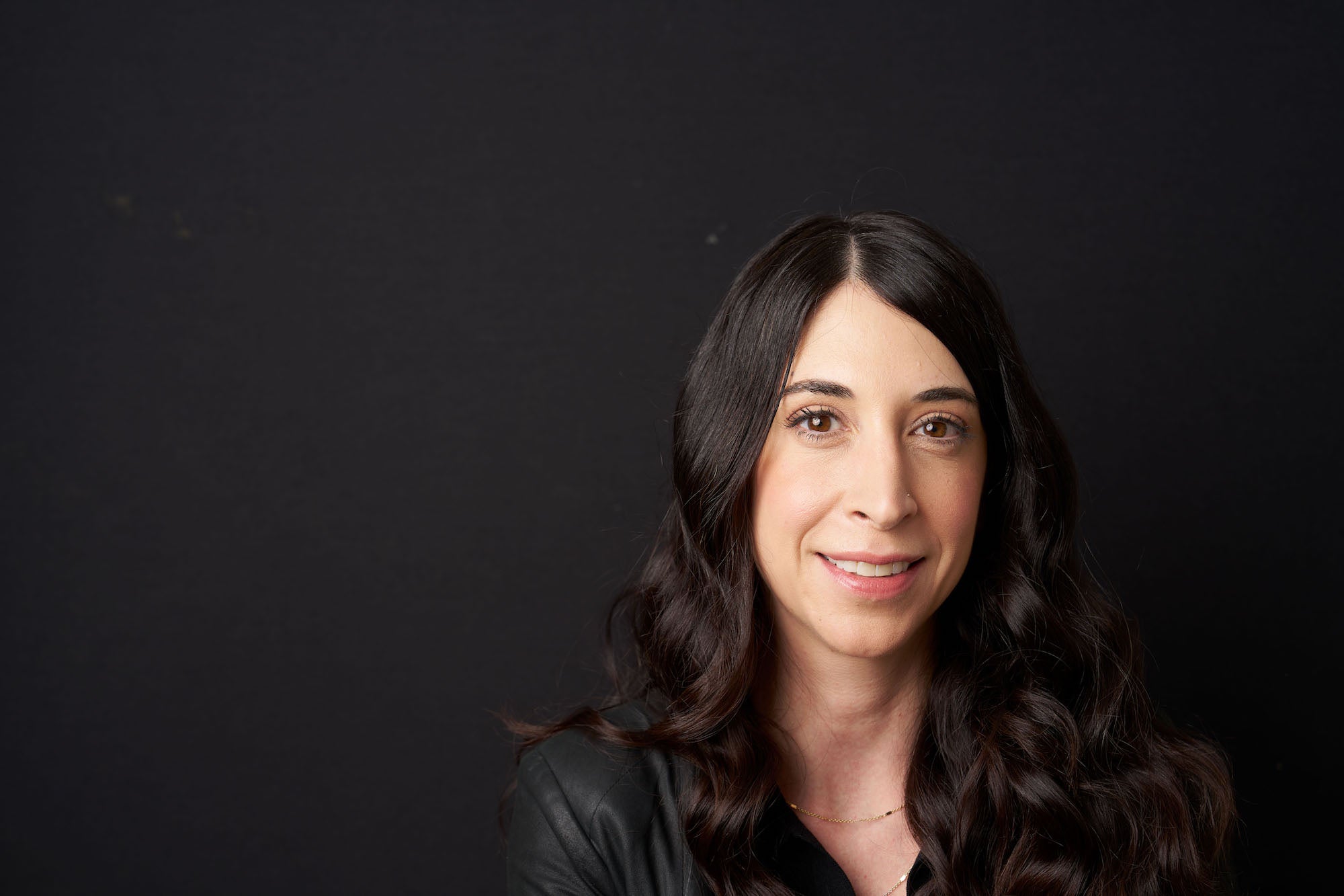 Studio headshot of a woman with long dark wavy hair against a black backdrop, softly lit and smiling.