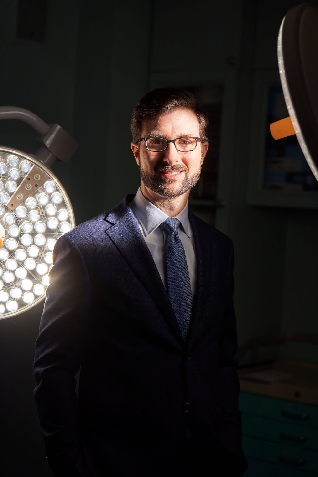 Professional portrait of a man in a suit and glasses lit by circular surgical lights in a clinical setting.