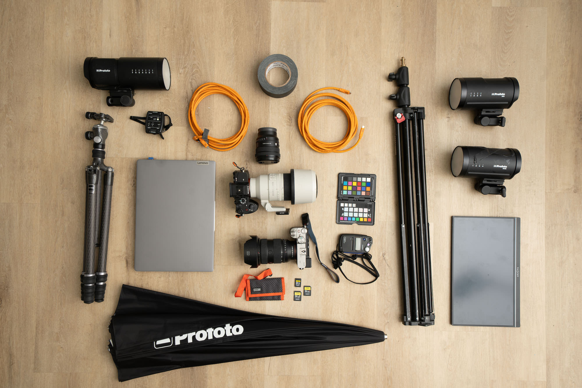 Flat lay of a headshot kit: Sony cameras and lenses, Profoto strobes, tripod, light stands, tether cables, laptop, monitor, color checker, cards, tape, and meter.