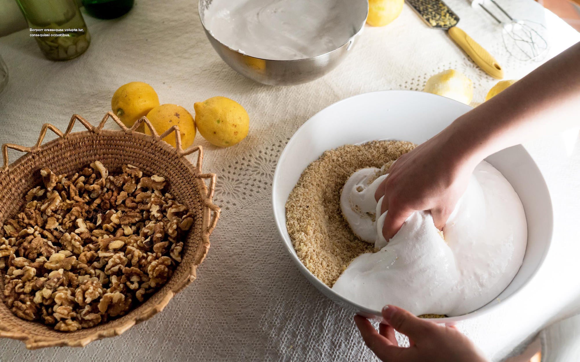 Hands fold fluffy meringue into ground nuts beside lemons and a basket of walnuts on a kitchen table. Hands fold fluffy meringue into ground nuts beside lemons and a basket of walnuts on a kitchen table.