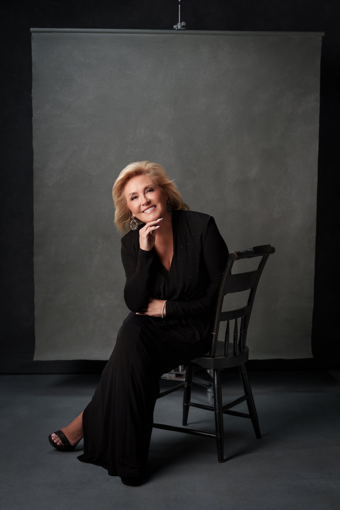Studio portrait of a woman in a black outfit seated on a chair, smiling with chin rested on hand against a gray backdrop. Studio portrait of a woman in a black outfit seated on a chair, smiling with chin rested on hand against a gray backdrop.